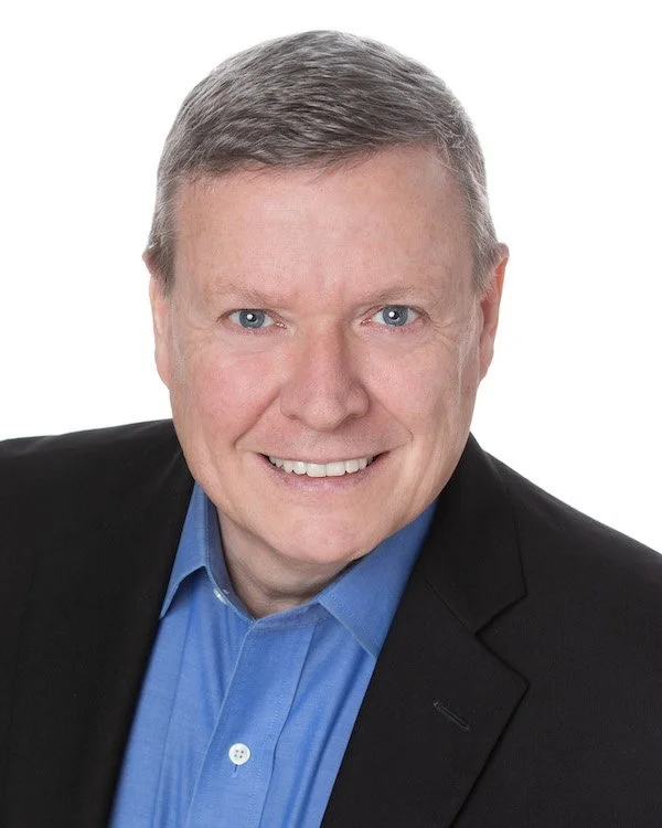 A middle-aged man with light gray hair, blue eyes, wearing a black blazer and a blue collared shirt, smiling against a white background.