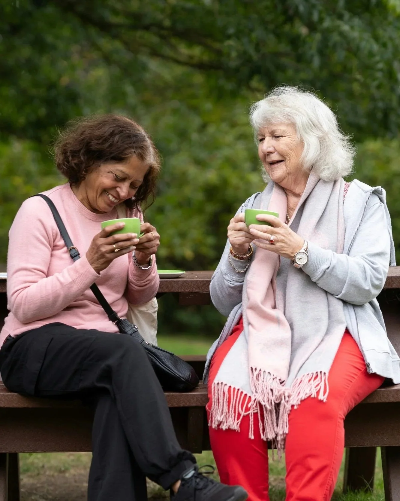 Two women sitting on a park bench, laughing and holding green cups, surrounded by green trees.