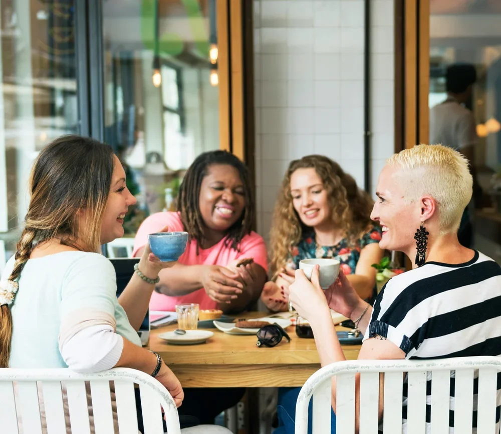 Group of diverse women sitting around a table, smiling, enjoying coffee and conversation in a cozy café setting.