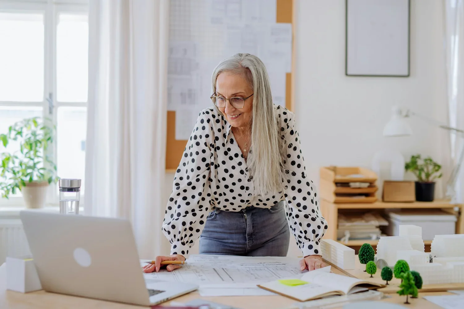 A woman with long gray hair and glasses wearing a polka dot shirt stands at a desk, looking at architectural plans or drawings. There are scale models of trees and building structures on the table. The room has large windows with natural light, light-colored walls, and a bulletin board.