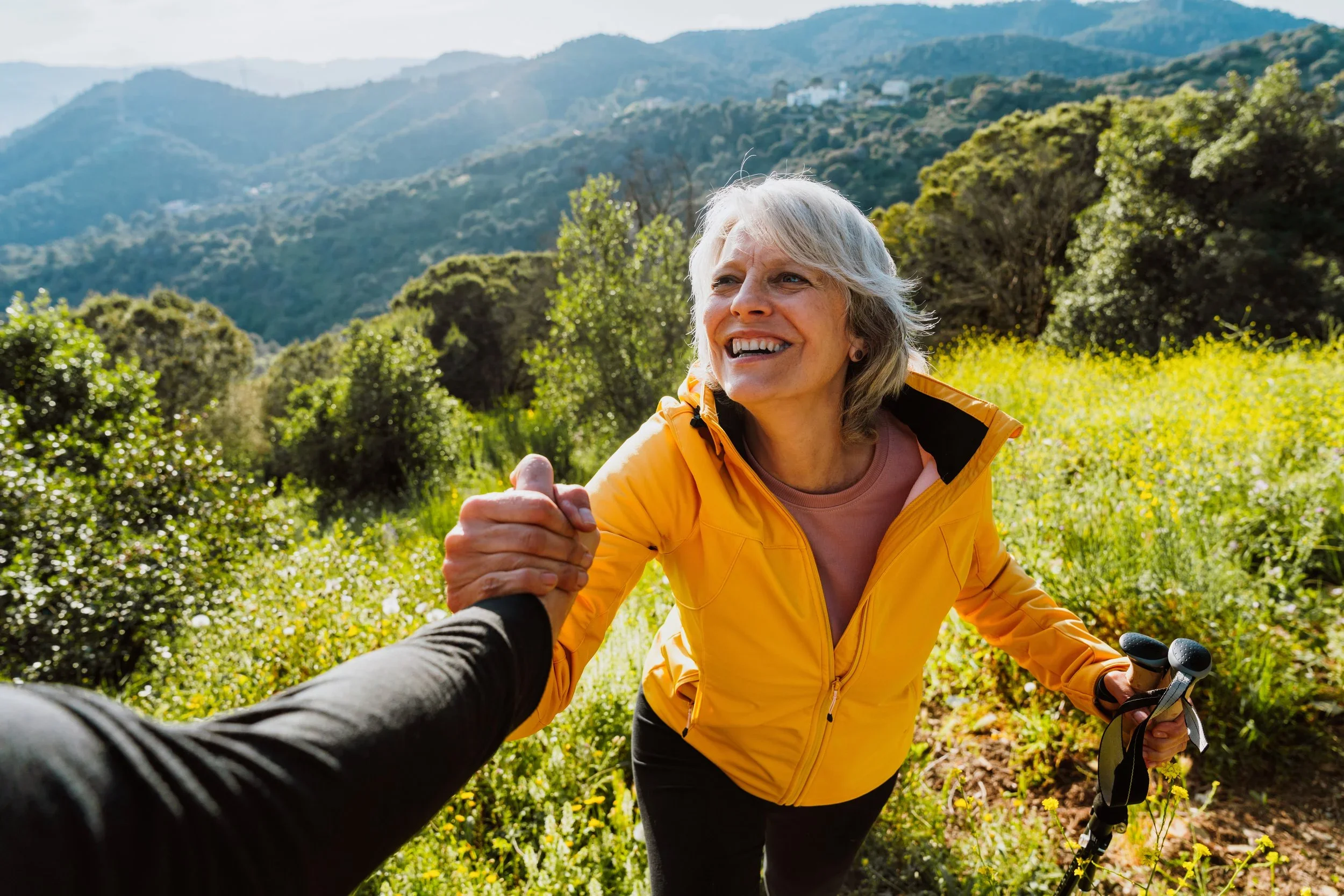 A smiling woman in a yellow jacket holding a person's hand outdoors in a lush green mountainous landscape.