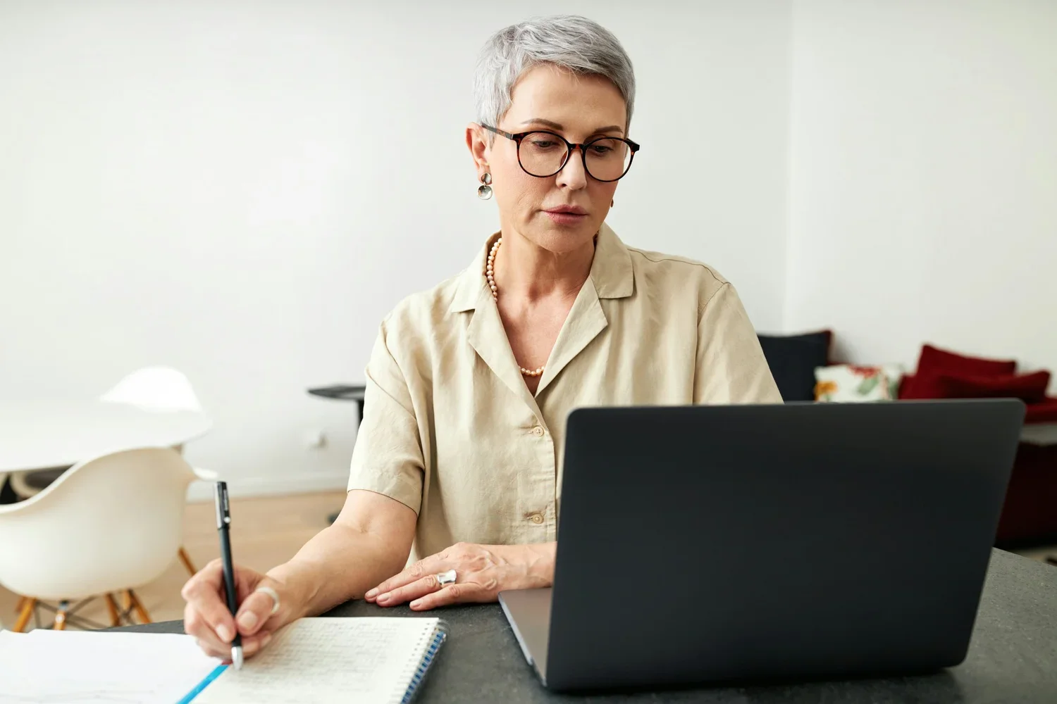 A woman with short gray hair, glasses, and jewelry, working on a laptop at a desk, taking notes in a notebook.