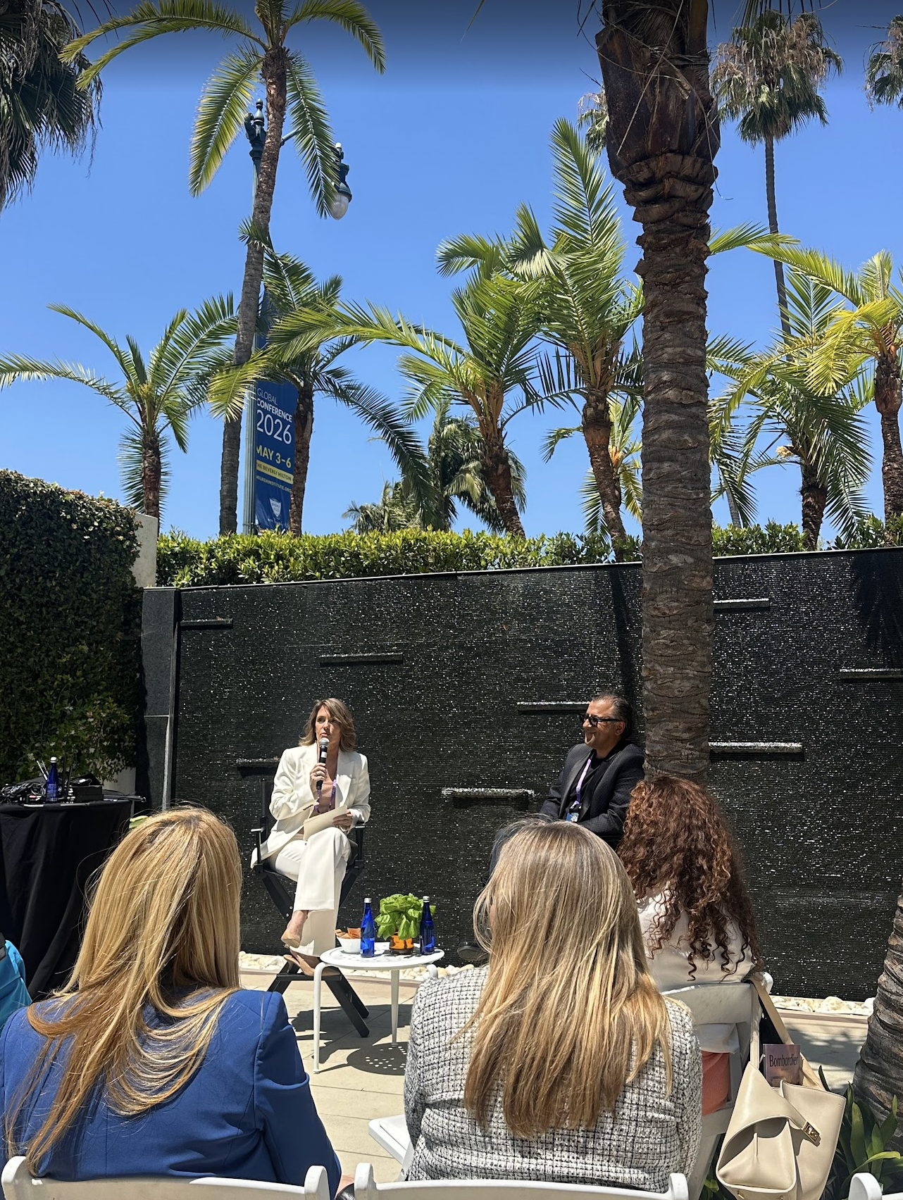 Outdoor conference with a woman in white speaking into a microphone and a man in glasses sitting next to her, surrounded by an audience seated at tables, with palm trees and a banner in the background.
