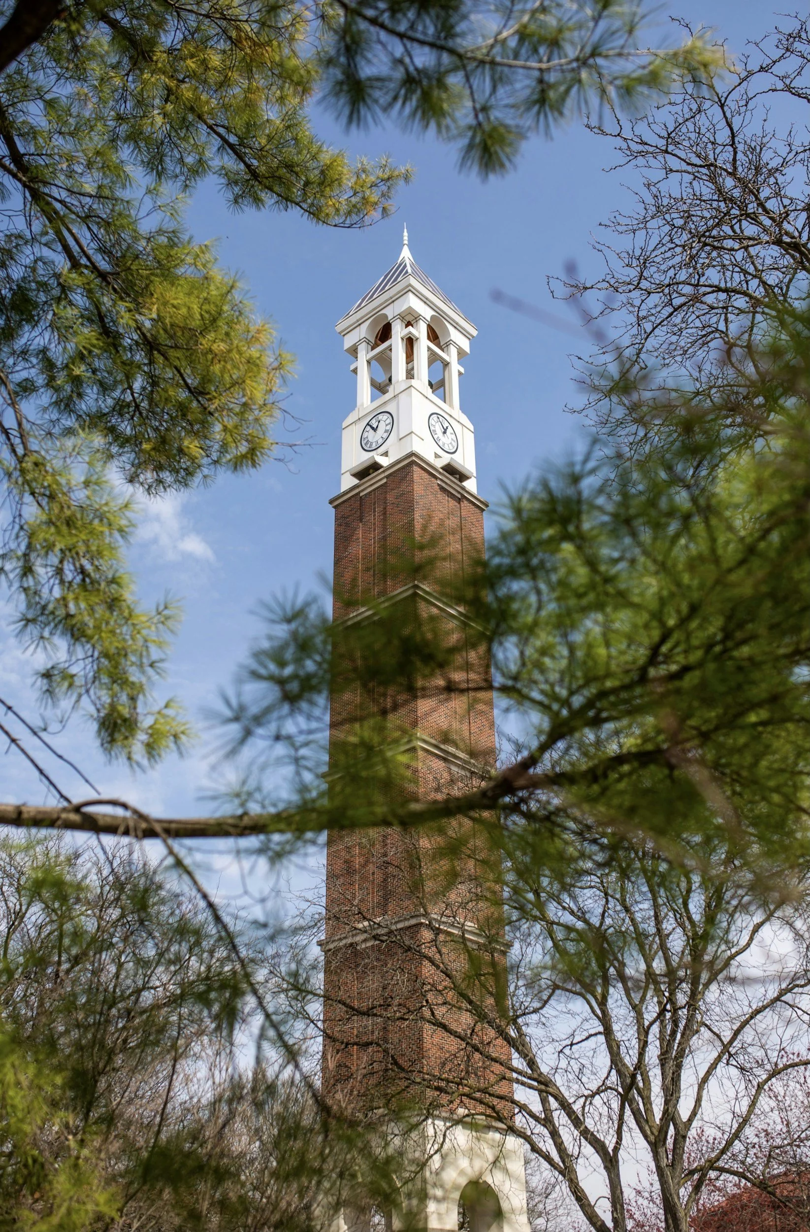 A clock tower with a white top and brick base, seen through trees with budding and leafless branches, against a partly cloudy blue sky.