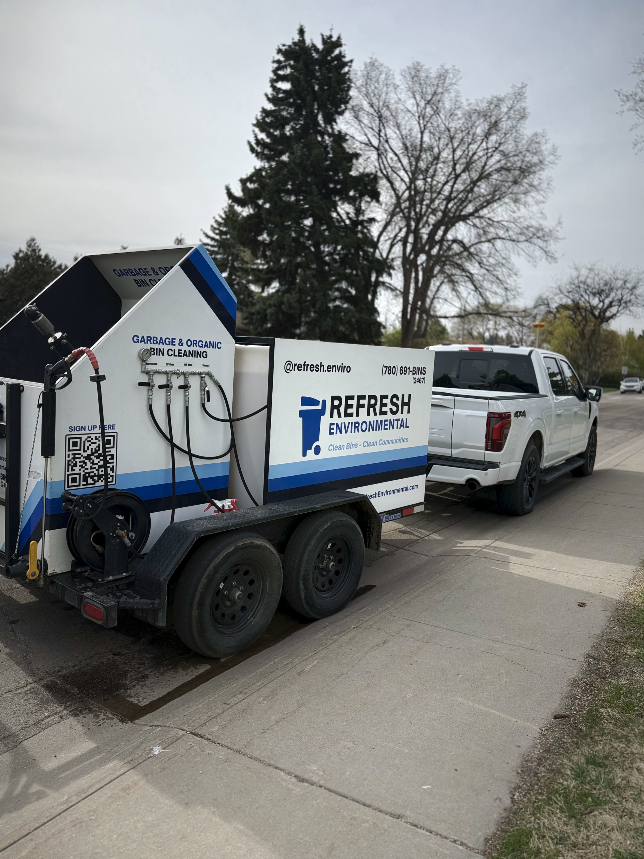A white pickup truck attached to a trash bin cleaning trailer labeled 'Refresh Environmental' parked on a parking lot. The trailer has hoses and equipment for trash bin cleaning.