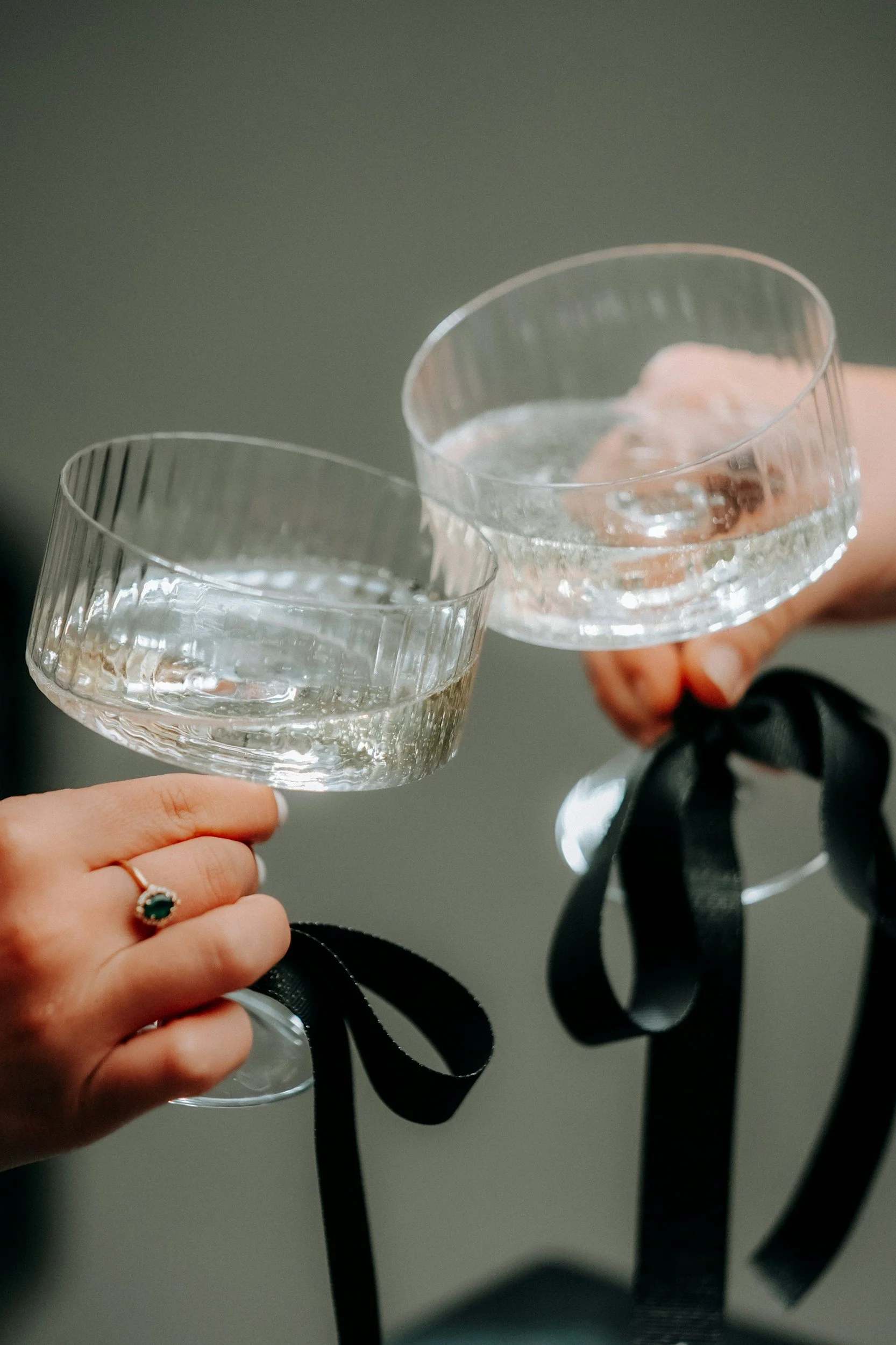 Two people holding champagne glasses with their hands visible, one with a ring, against a plain background.