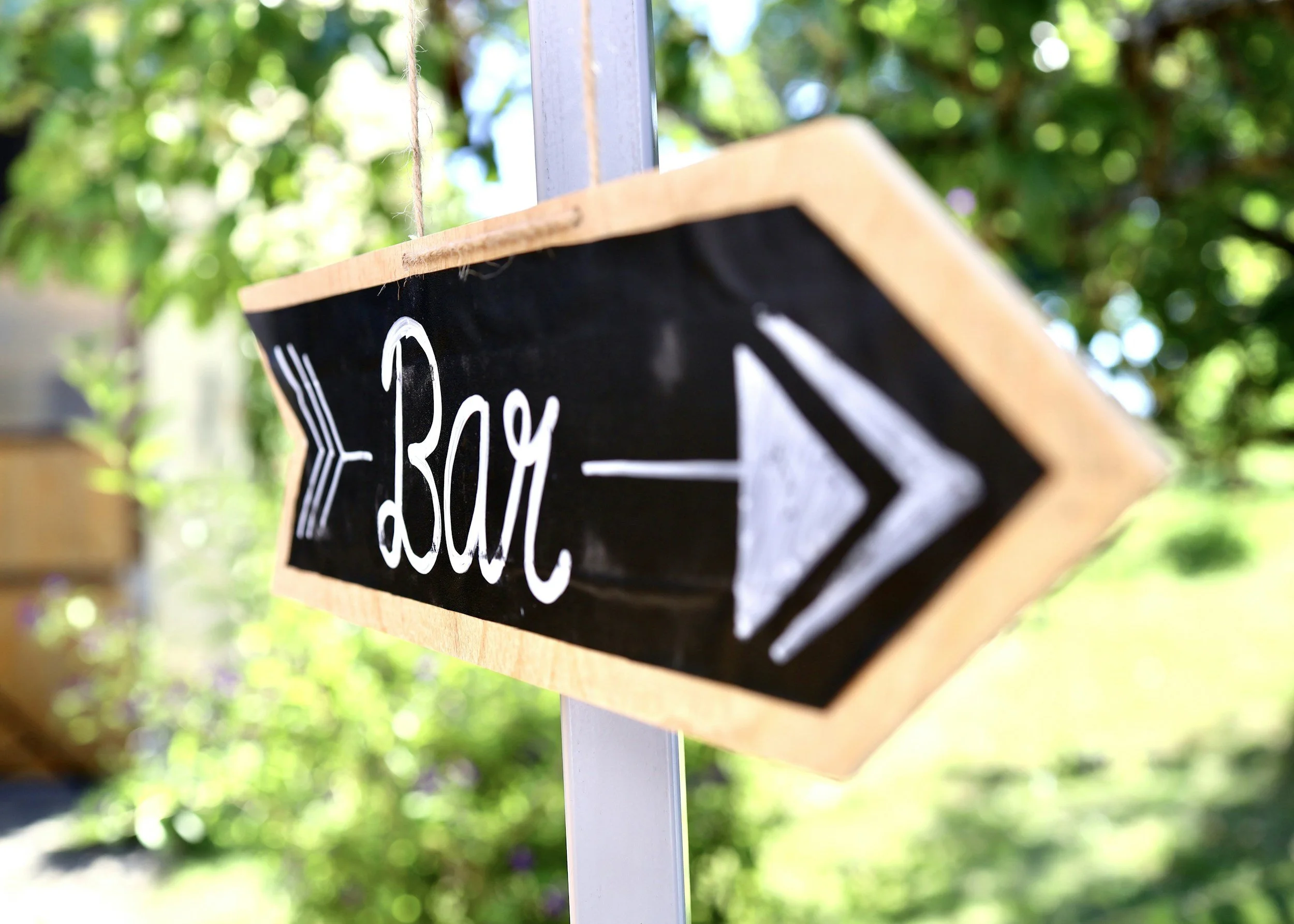 A black chalkboard sign with white handwritten text that says 'Slow' is hanging from a wooden frame with a metal chain, outdoors surrounded by greenery and sunlight.
