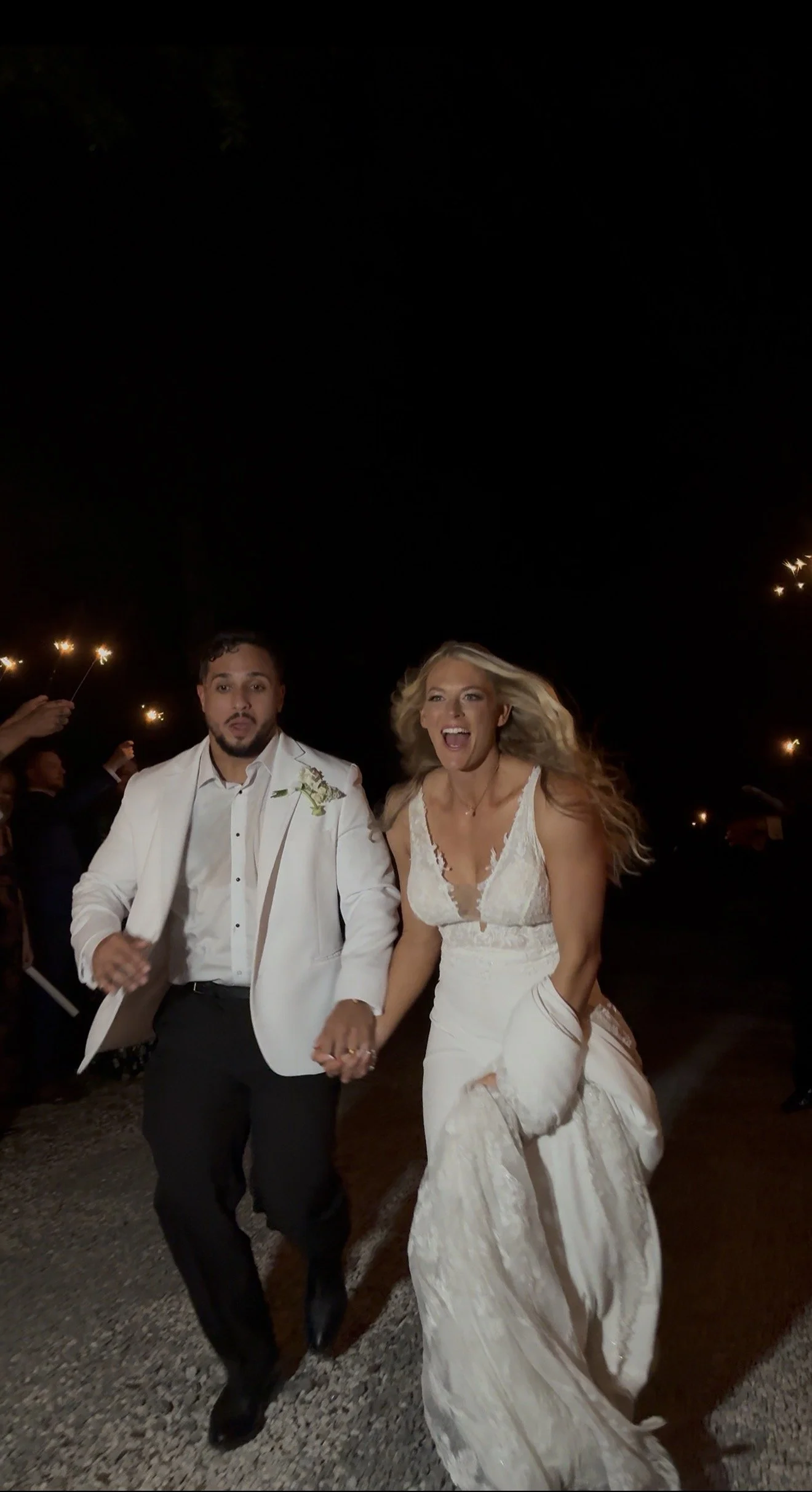 A couple dressed in wedding attire running hand in hand at night, with people holding sparklers in the background.
