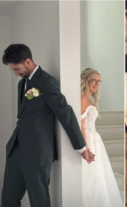 A bride and groom are standing back to back, holding hands but not looking at each other, in a wedding venue.