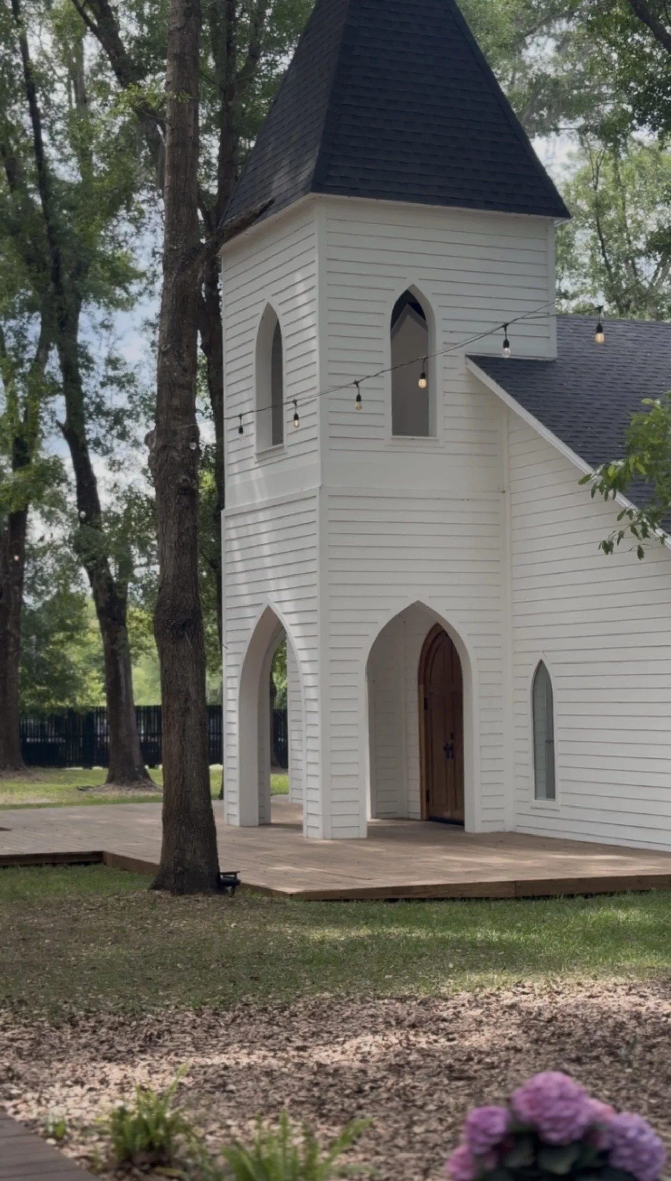 A small white church (Whitewood Ranch, in Florida) with a black roof and arched windows, set in a wooded area with hanging string lights and purple flowers in the foreground.