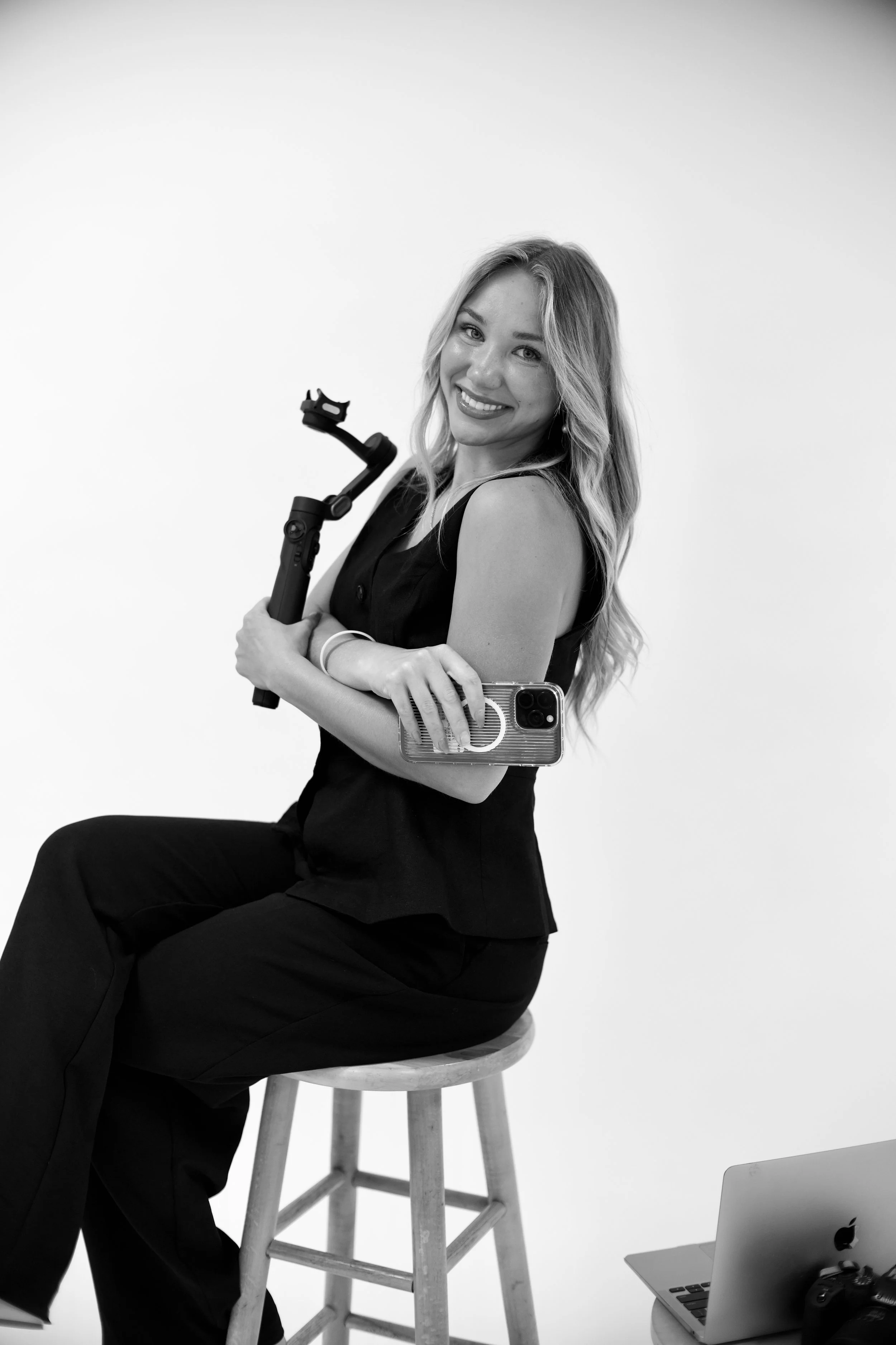 A smiling woman with long wavy hair, wearing a sleeveless top, sitting on a stool holding a camera. She has a phone in a clear case attached to her waist, and there is a laptop and camera on a table nearby. The background is plain and white.
