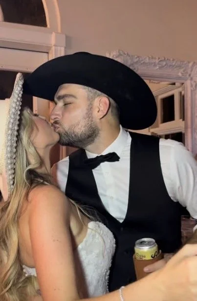A man and woman dress as a cowboy and cowgirl sharing a kiss at a celebration or wedding reception.