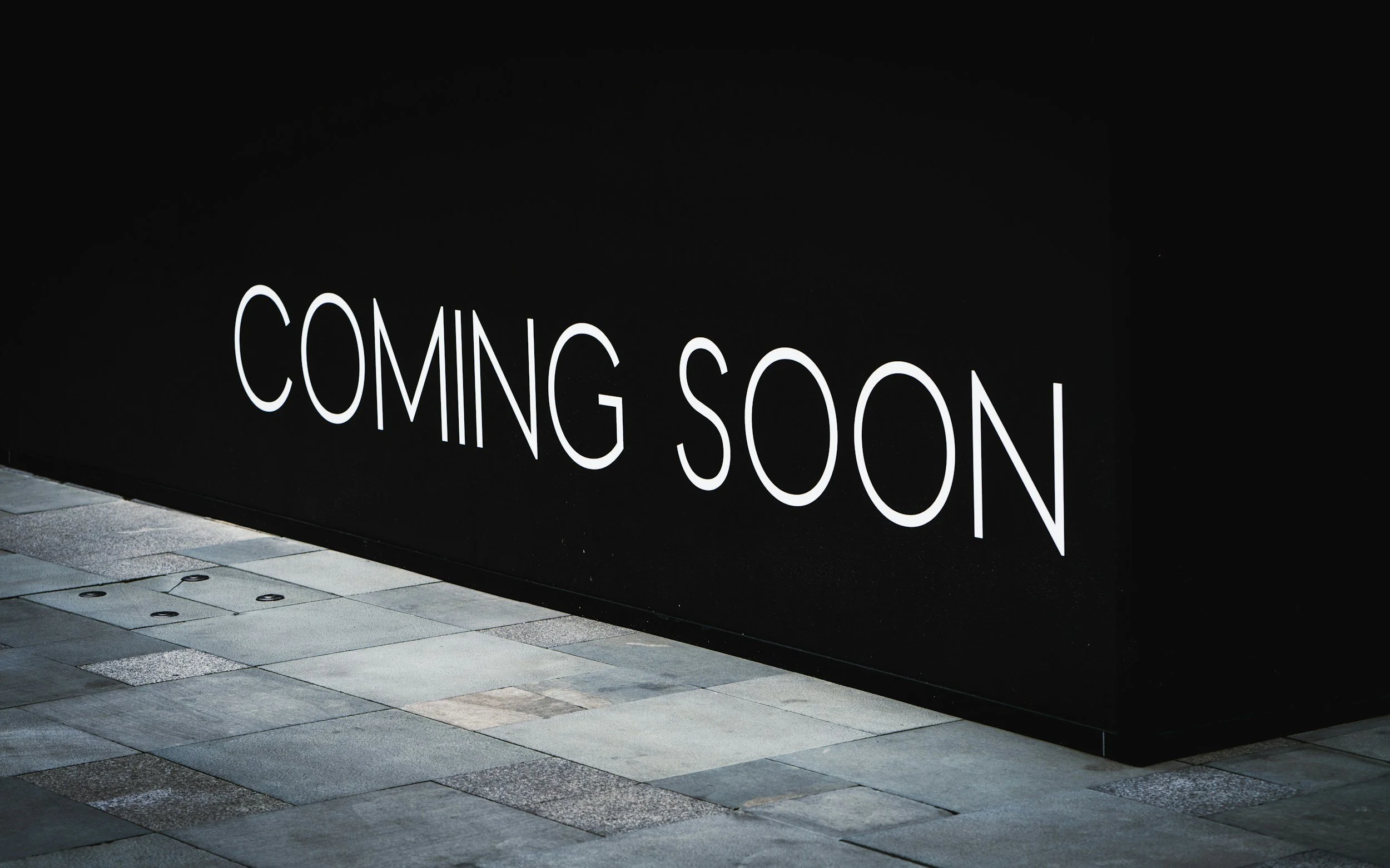 A black wall with illuminated white text that reads 'COMING SOON' and a sidewalk in front of it.