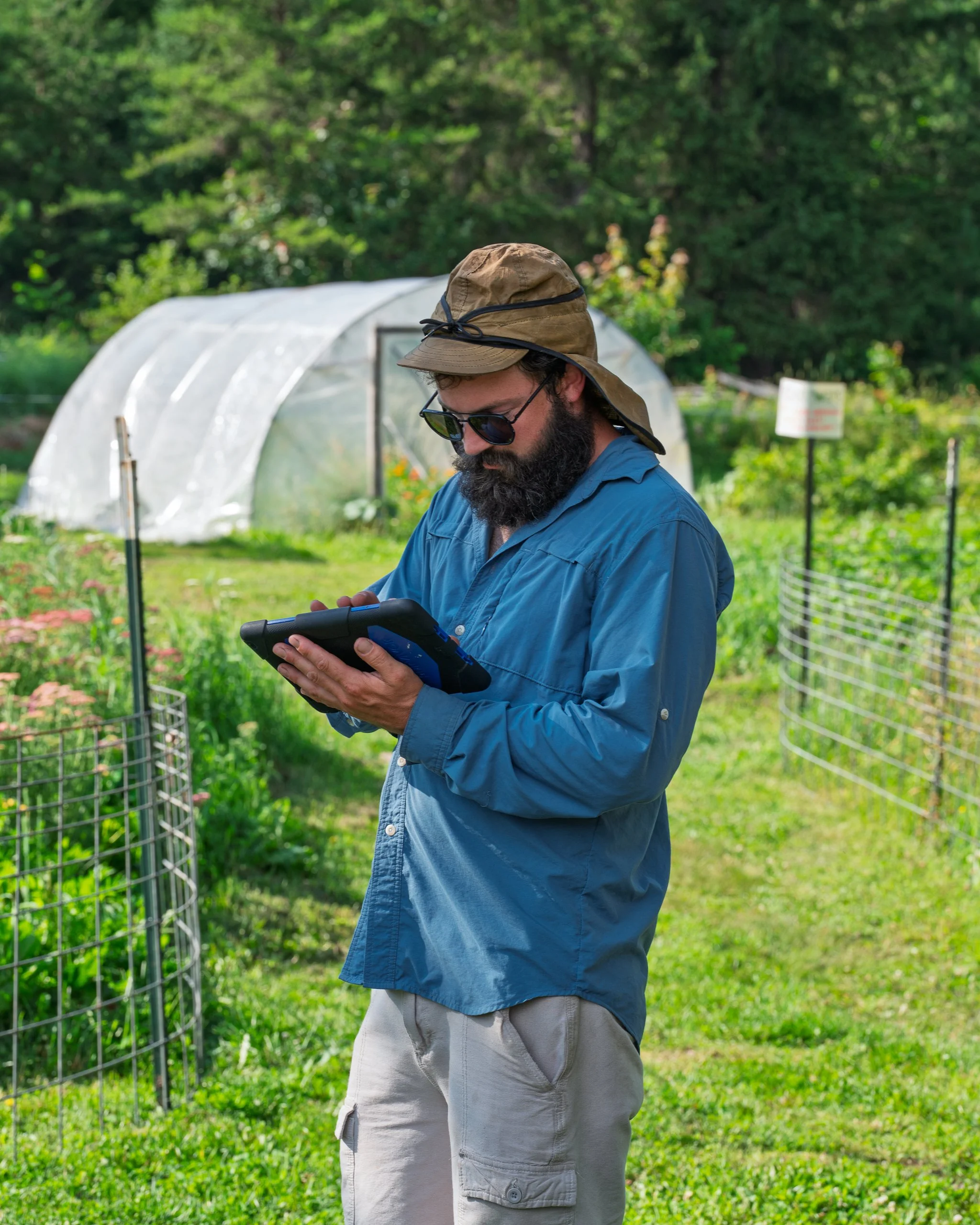 A man with a beard wearing sunglasses, a wide-brimmed hat, a blue shirt, and beige shorts standing outdoors in a garden or farm with a greenhouse and greenery in the background.