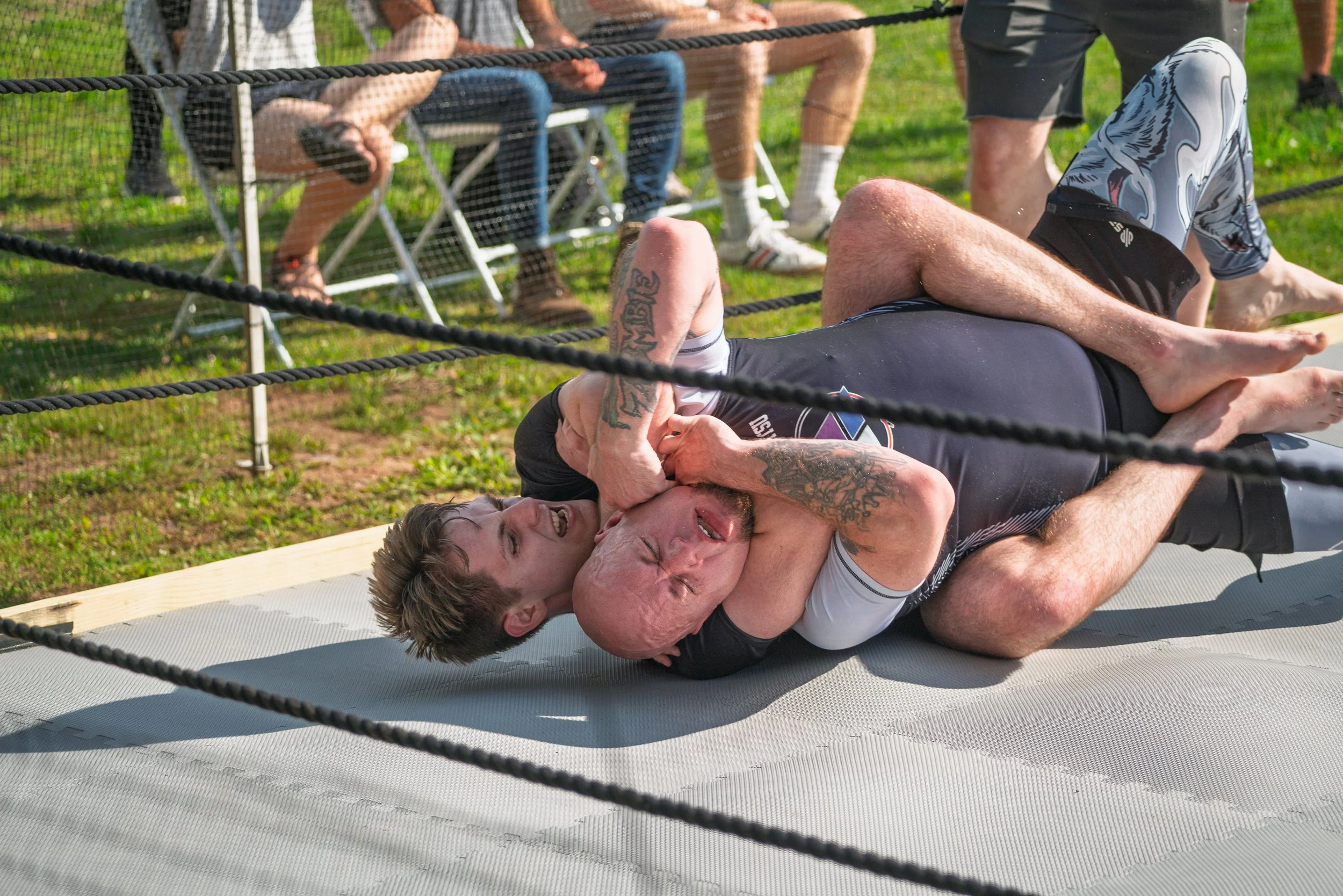 Two men are wrestling on a mat outdoors, surrounded by a rope barrier, with three people sitting on chairs watching in the background.