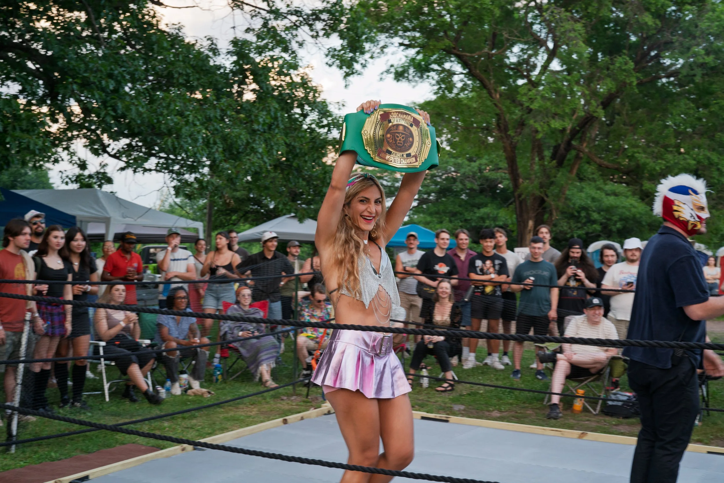 A woman with long, wavy blonde hair wearing a ritual dance costume and holding a championship belt above her head in a boxing ring at an outdoor event with a crowd of spectators and trees in the background.
