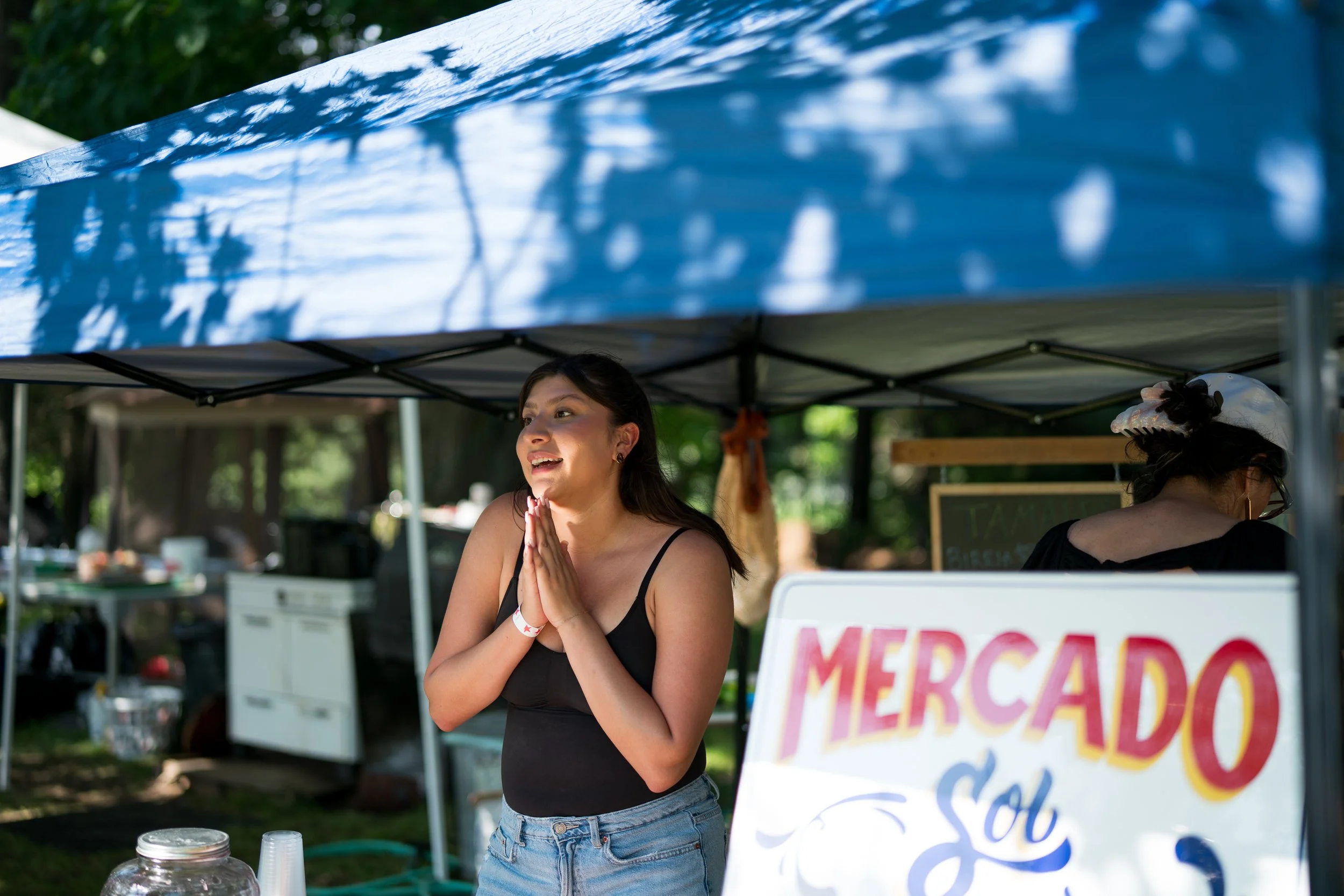 A young woman wearing a black dress and blue jeans, standing under a blue canopy, with her hands pressed together in front of her chest, smiling. There is a cake jar and plastic cups on a table in front of her, and a partial sign reading 'MED' and 'S