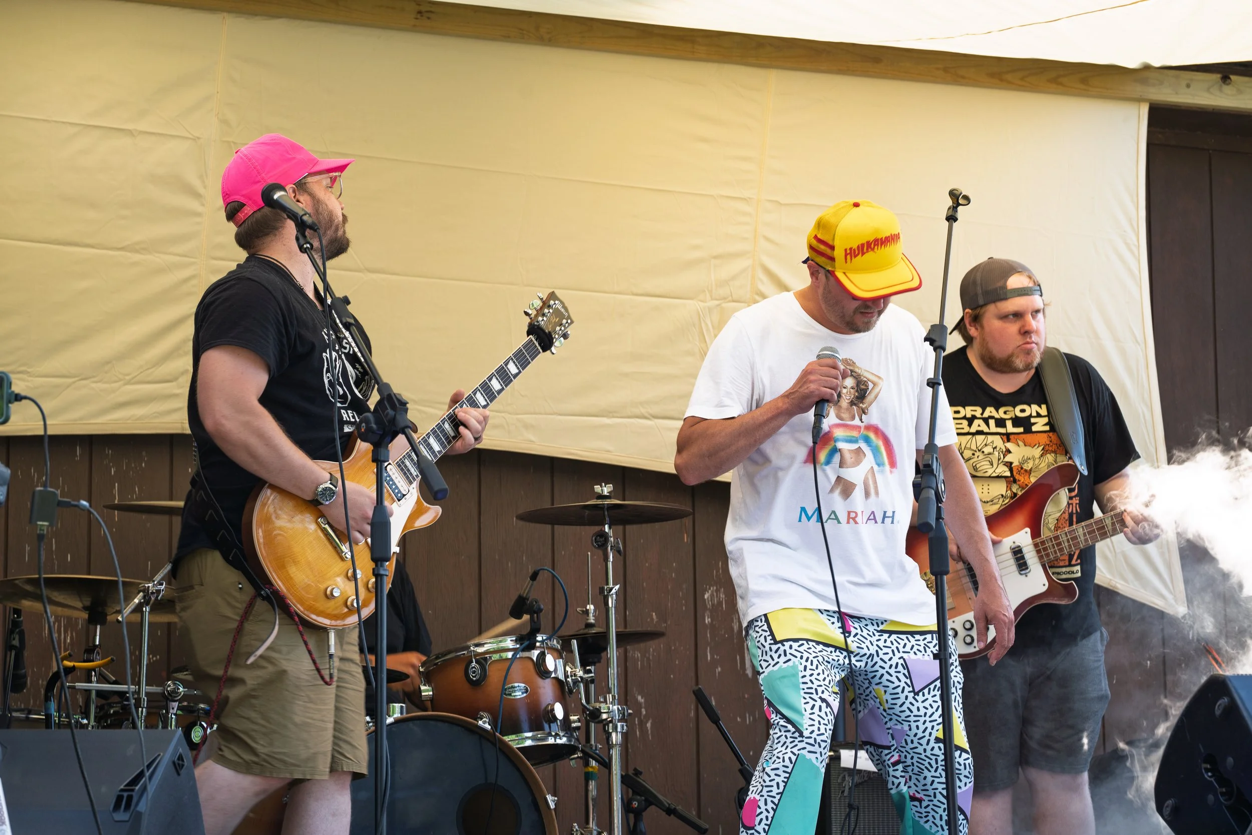 Three men performing with guitars and microphone on a stage with yellow backdrop. One wears a pink baseball cap, another a yellow cap with red text, and the third a gray cap. One man is singing, and the others are playing guitar.