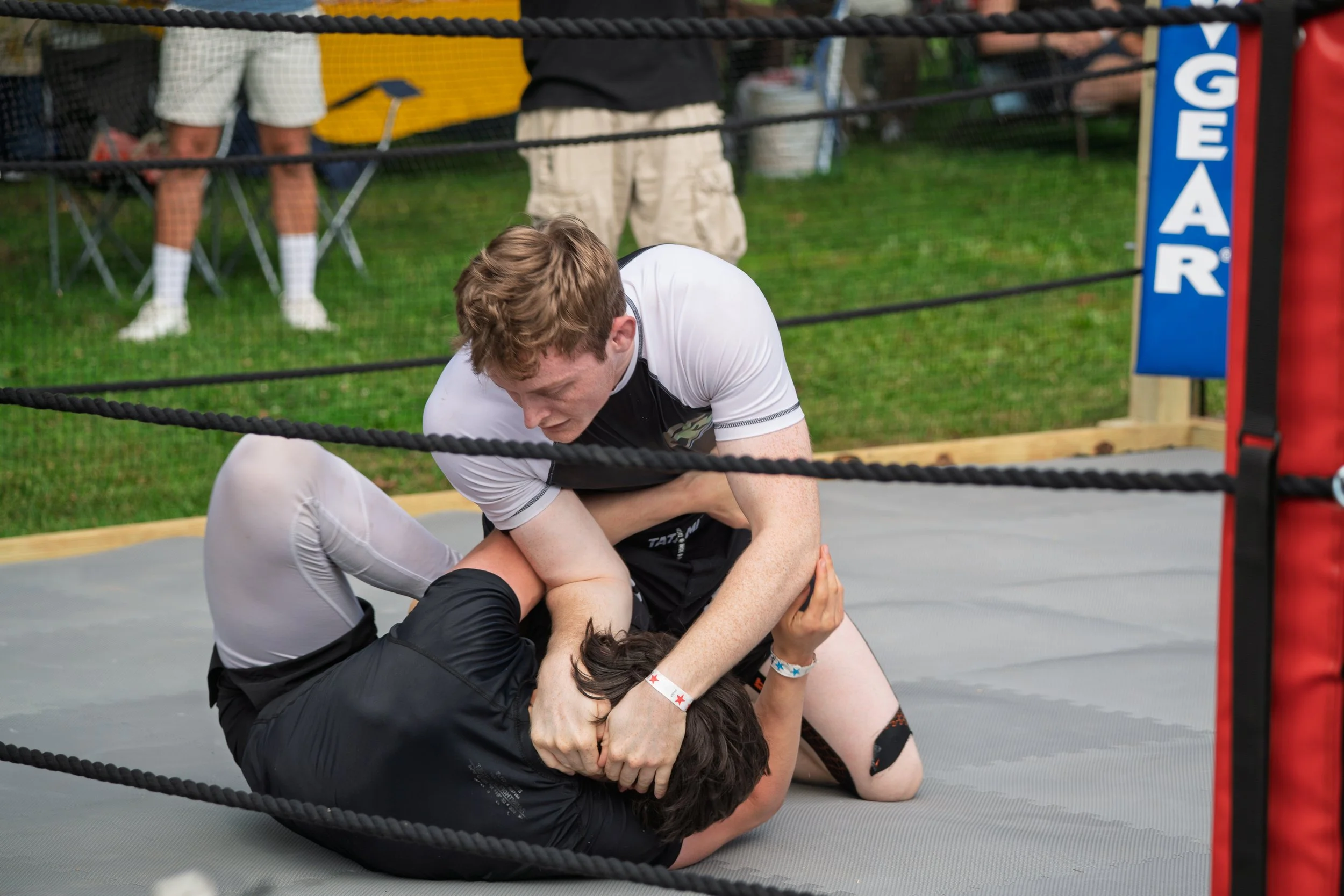 Two men are grappling on the ground inside a wrestling ring, while other people stand outside the ring watching. One man appears to be pinning the other.