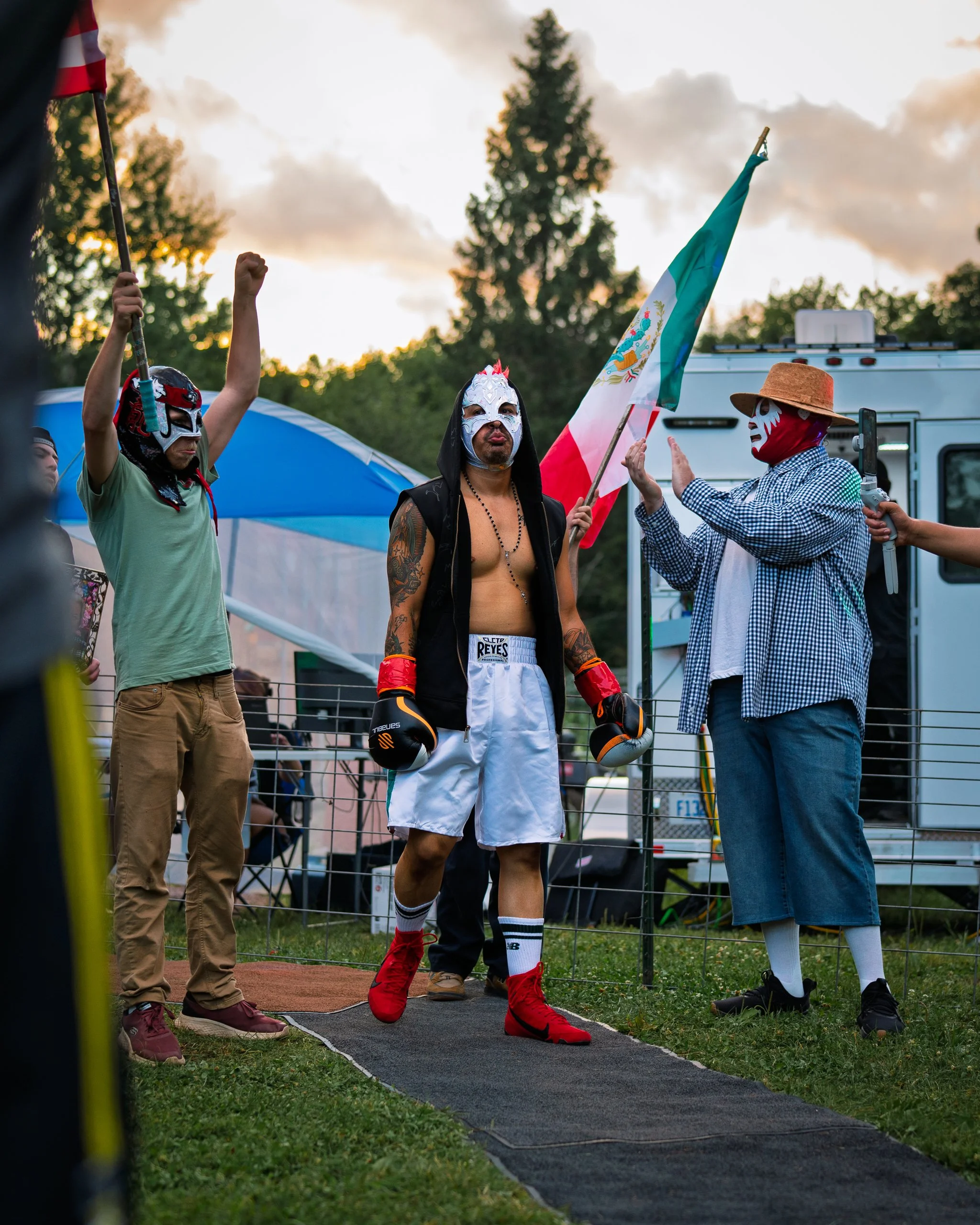 Boxer wearing a dragon mask, white shorts, red boxing gloves, and red shoes, standing at a boxing ring, with a man in a straw hat and mask holding a camera nearby, and people observing the scene outdoors at sunset.