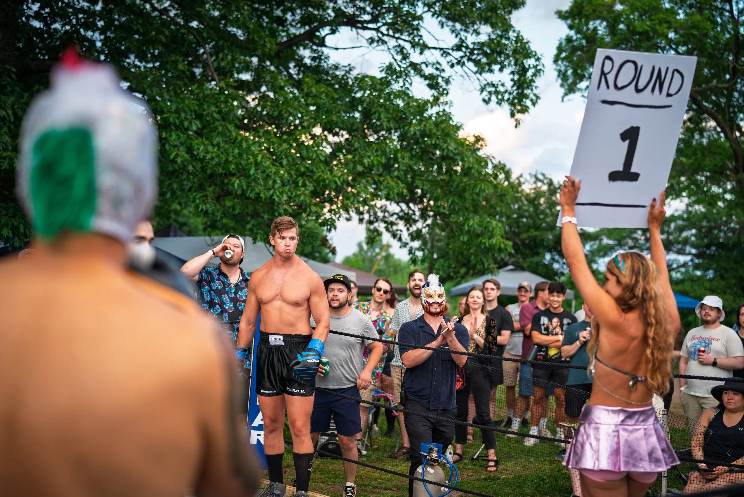 A crowd of people gathered outdoors, standing inside a boxing or wrestling ring with ropes. Some people are wearing masks, casual clothing, and sunglasses. One person holds a sign that says 'Round 1.' The background shows trees and an overcast sky.