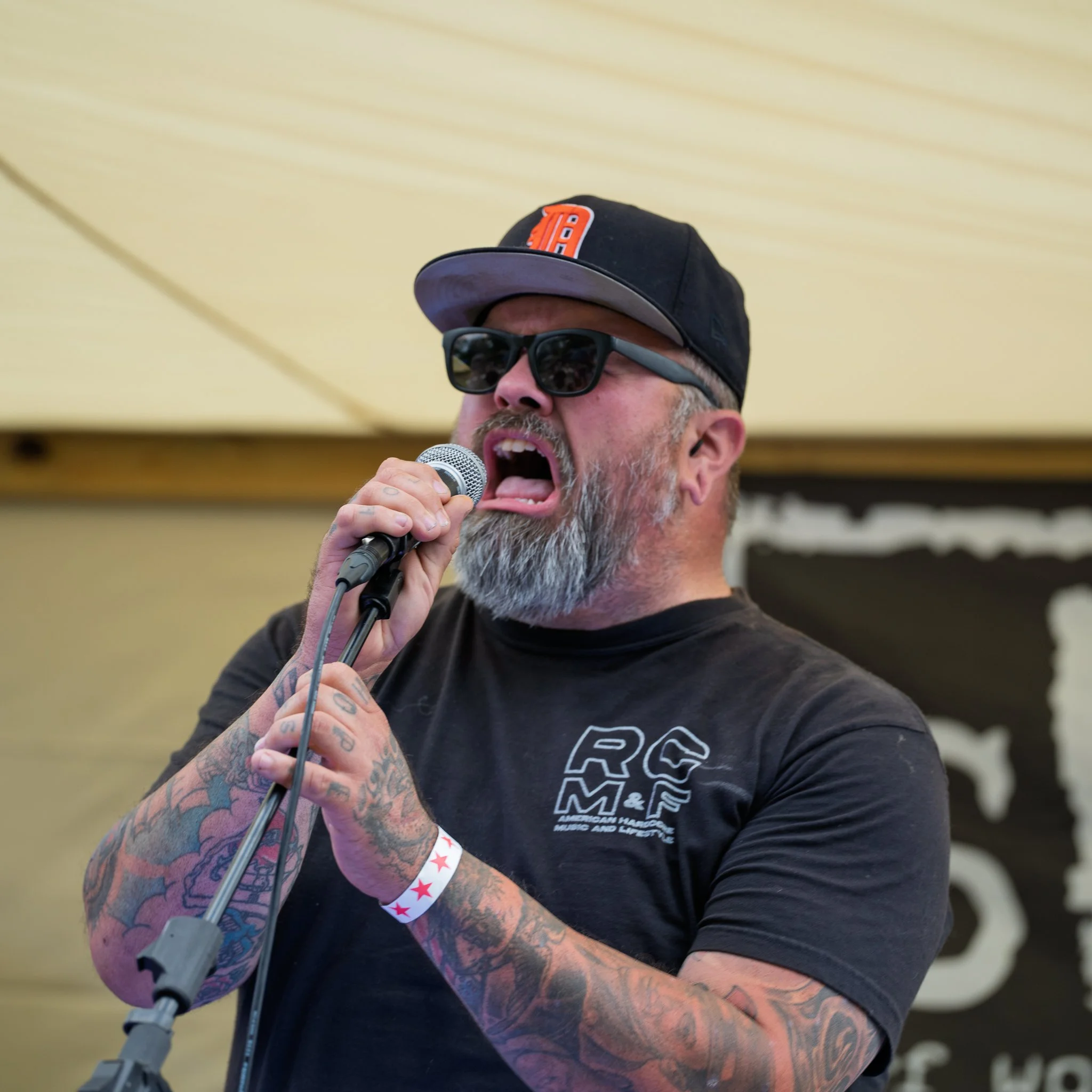 A man with tattoos on his arm singing into a microphone. He wears a black cap with an orange and white logo, black sunglasses, and a black t-shirt with white graphics.
