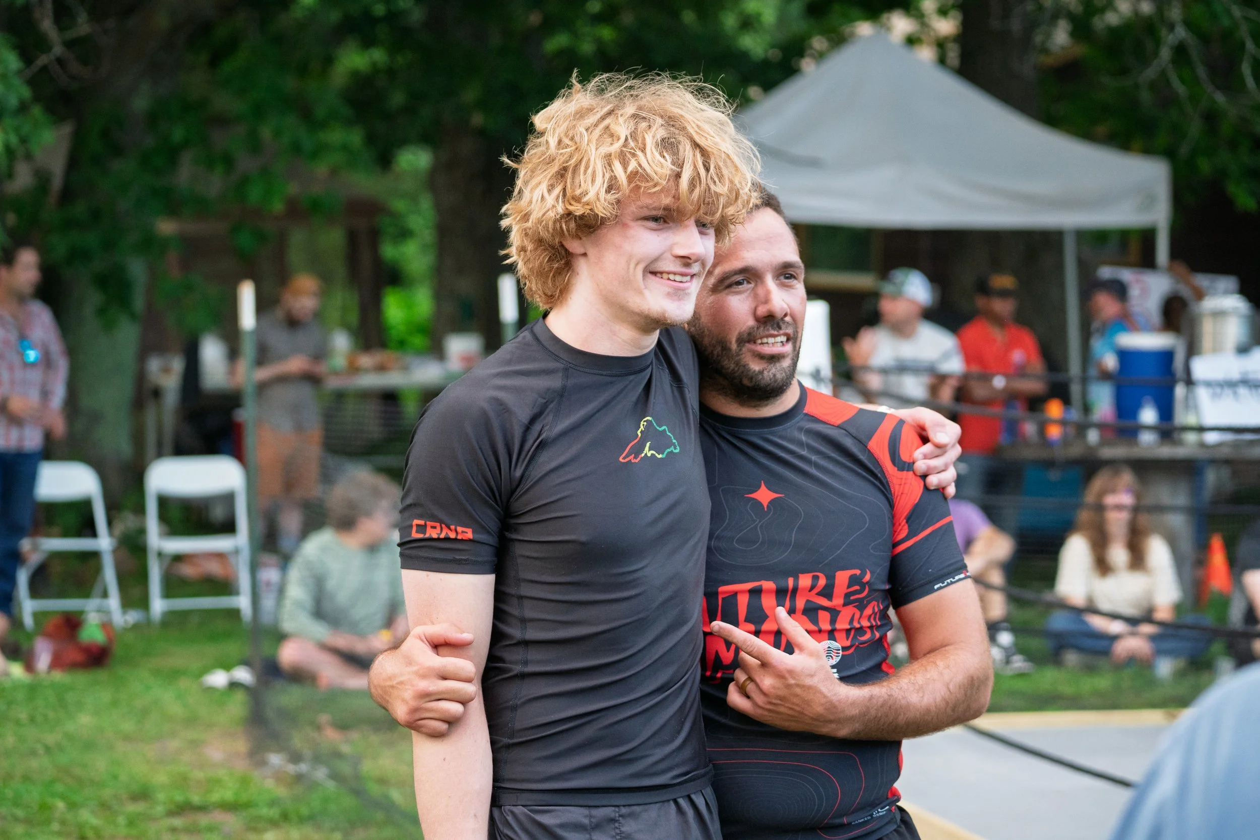 Two smiling young men with athletic build and curly hair embracing at an outdoor event, with trees, a white canopy tent, and people in the background.