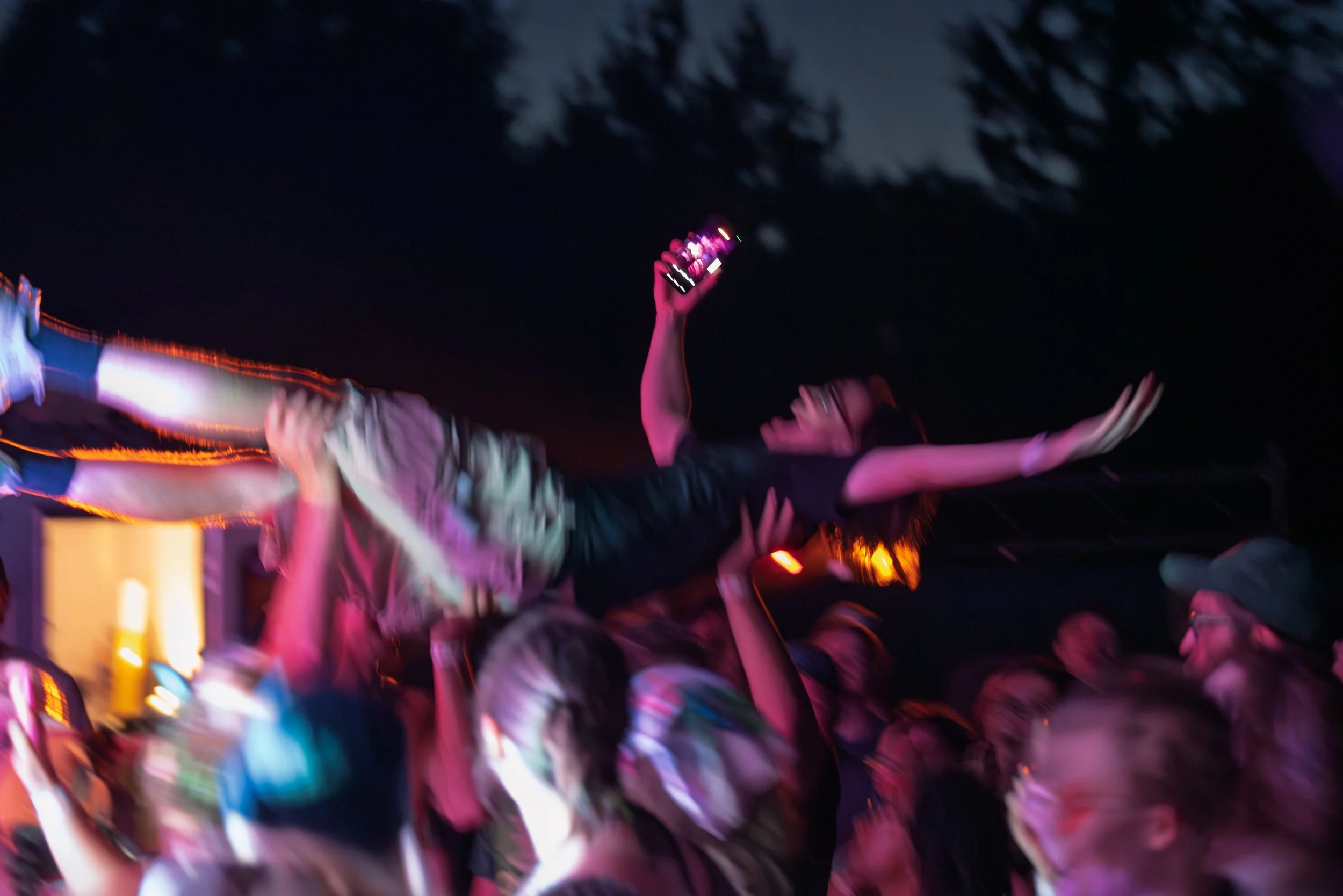 People crowd together outdoors at night, reaching up with smartphones to take pictures or videos, illuminated by colorful pink and orange lights.