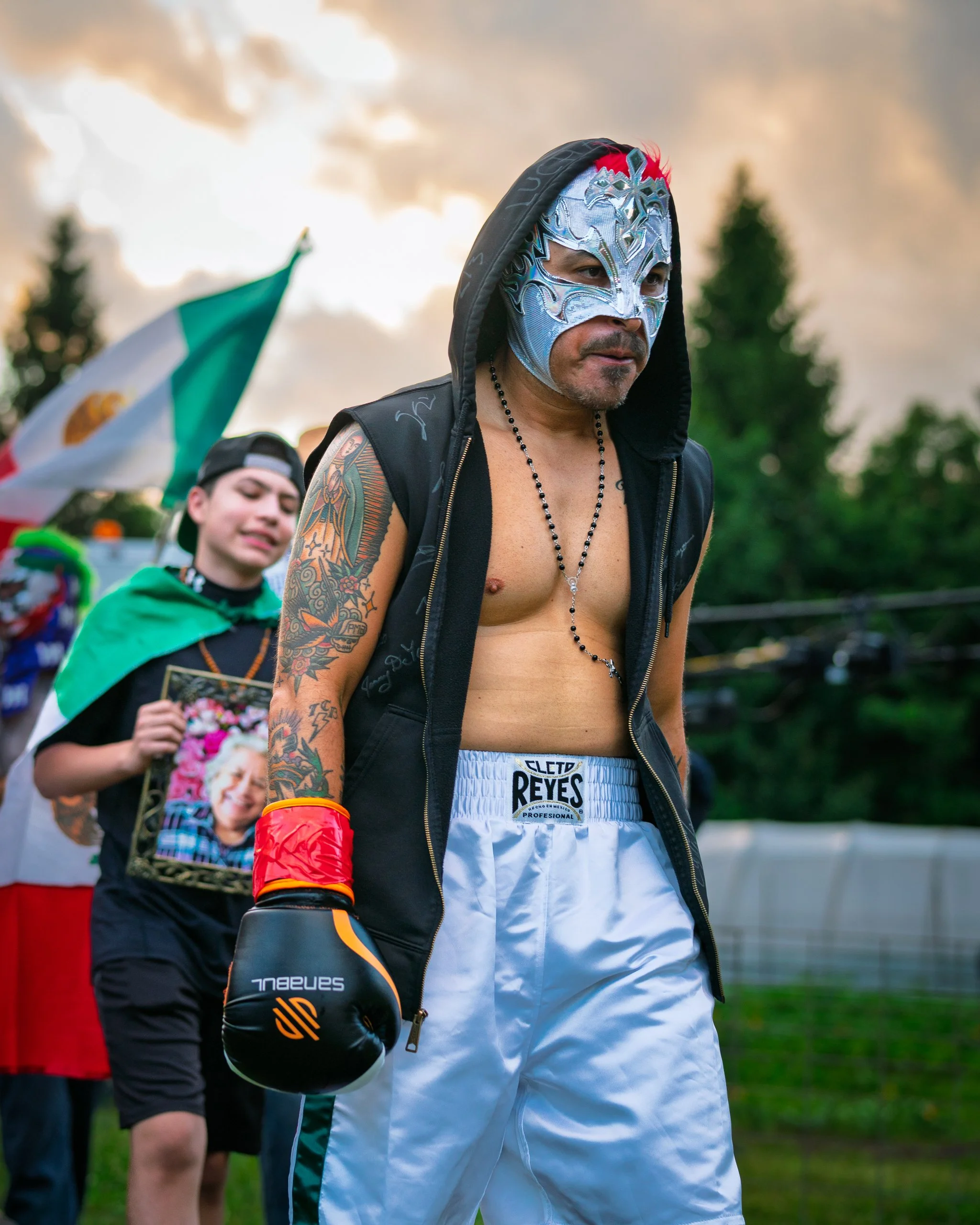 A male boxer wearing a silver lucha libre mask with red accents, a black hoodie, boxing trunks, and gloves, with tattoos on his arms, standing outdoors with a cloudy sky and trees in the background. A person with a Mexican flag and a Mickey Mouse pic
