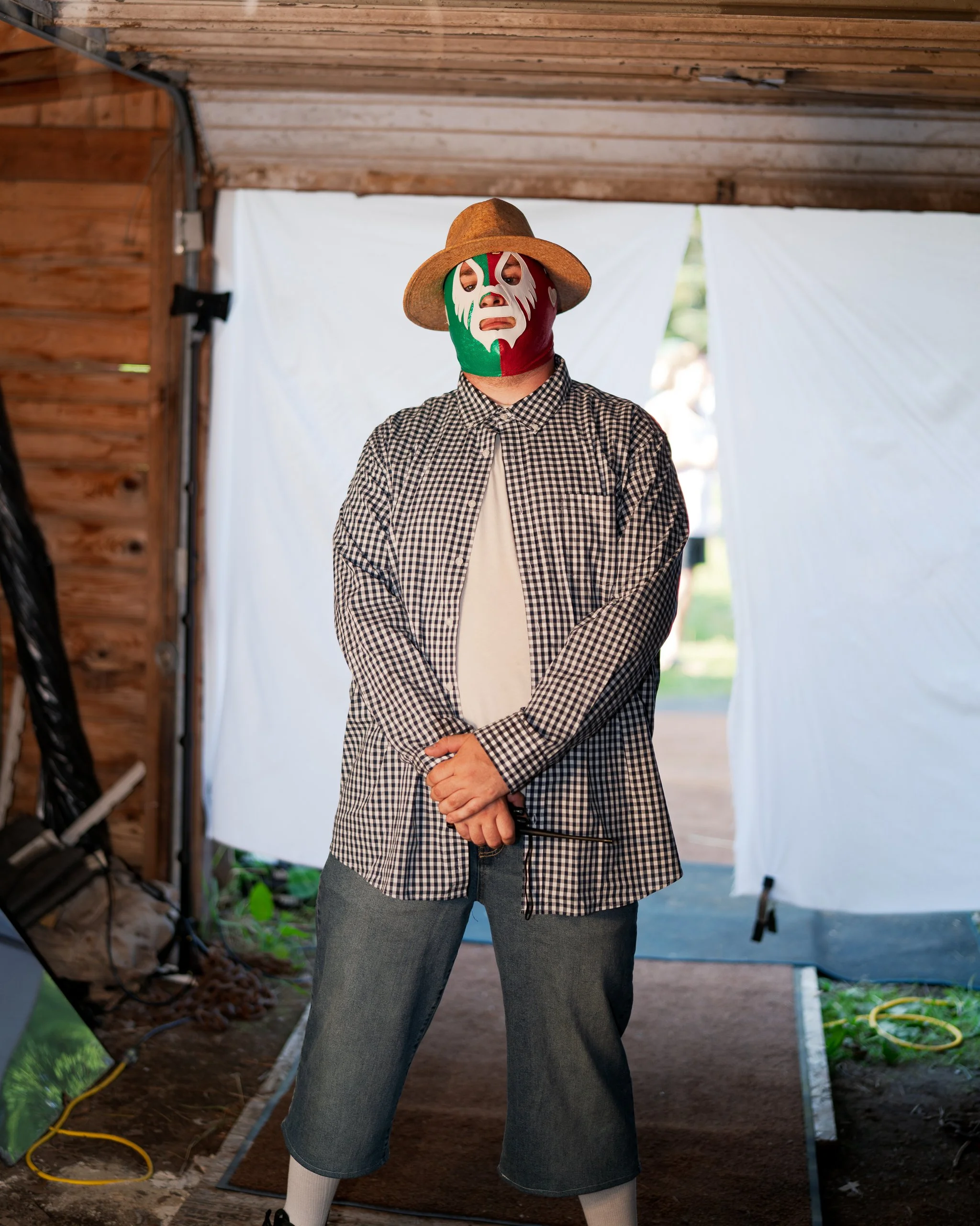 Individual wearing a lucha libre mask with Mexican flag colors, a straw hat, a checkered shirt, and denim shorts, standing inside a wooden and fabric structure.