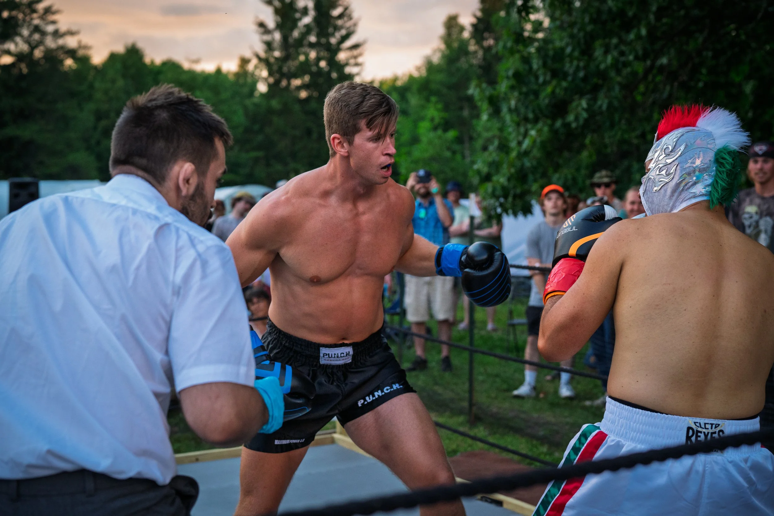 Two shirtless men and a man in a white shirt and gloves are engaged in a martial arts match outdoors, surrounded by a crowd of spectators with trees in the background.