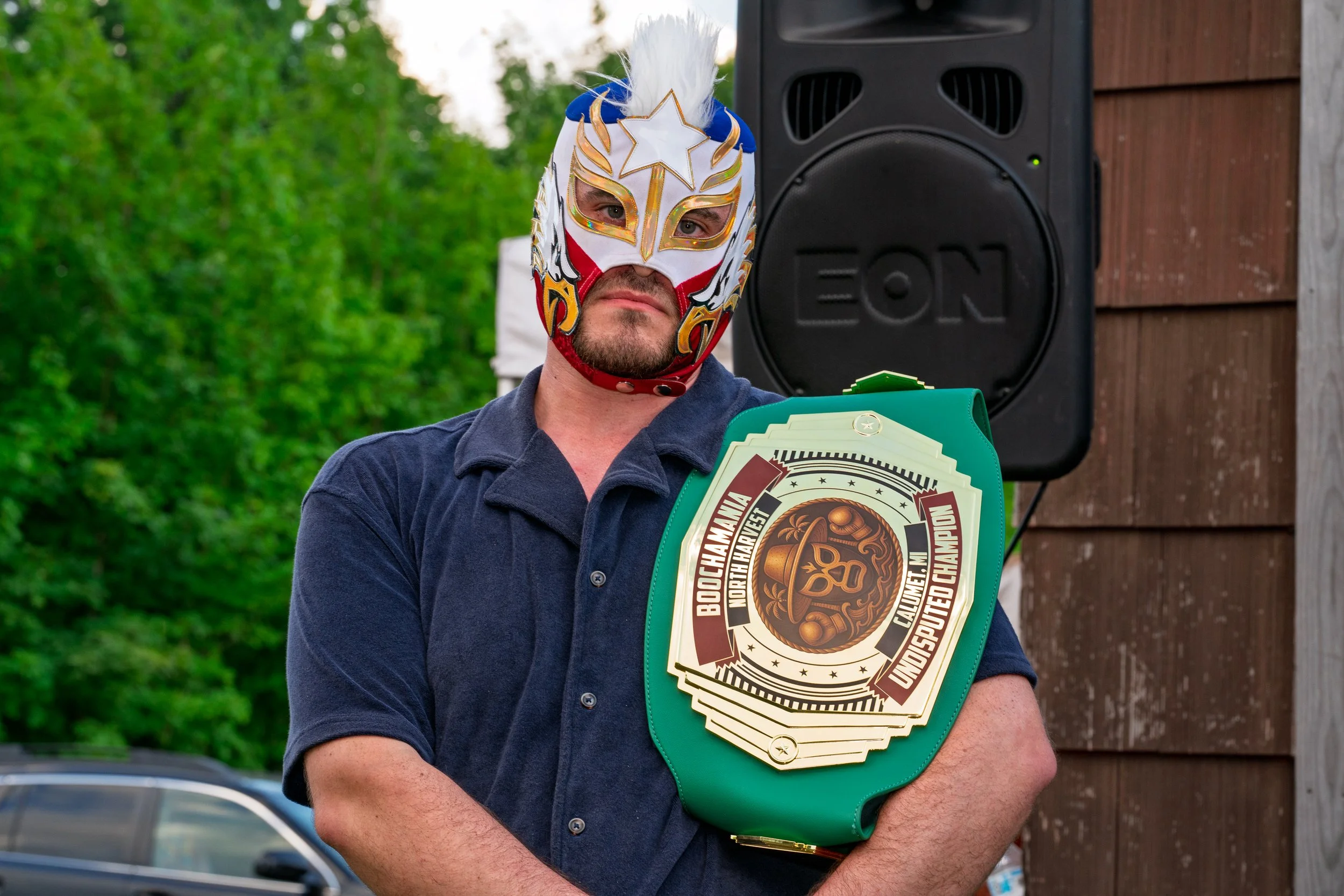 A man wearing a colorful luchador mask with a white and gold design and a blue top, standing outdoors near a large black speaker. He is holding a green championship belt with a gold and black emblem that reads "BOUCHAMANIA NORTH HARVEST UNIFIED DISP.