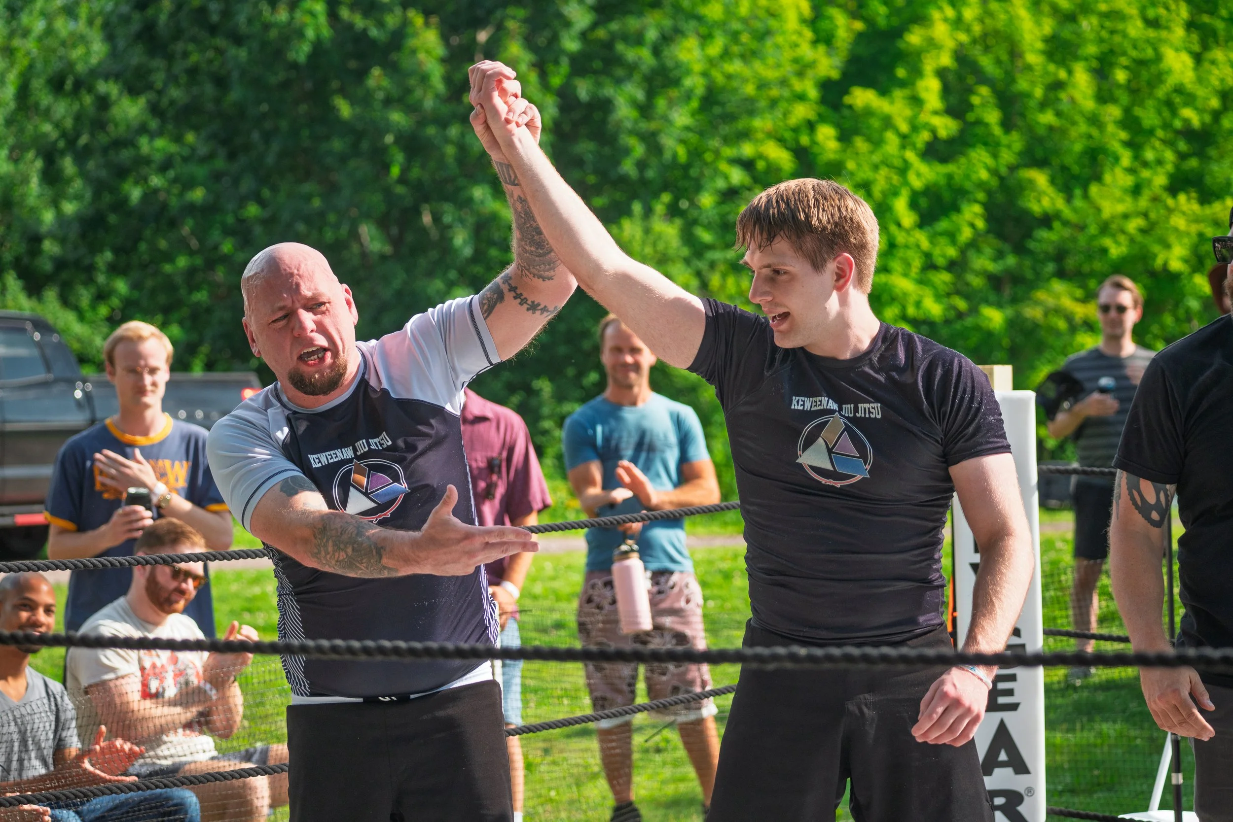 Two men are in a ring arguing or talking, with one arm raised. Several people are watching and cheering in the background, outdoors with green trees.