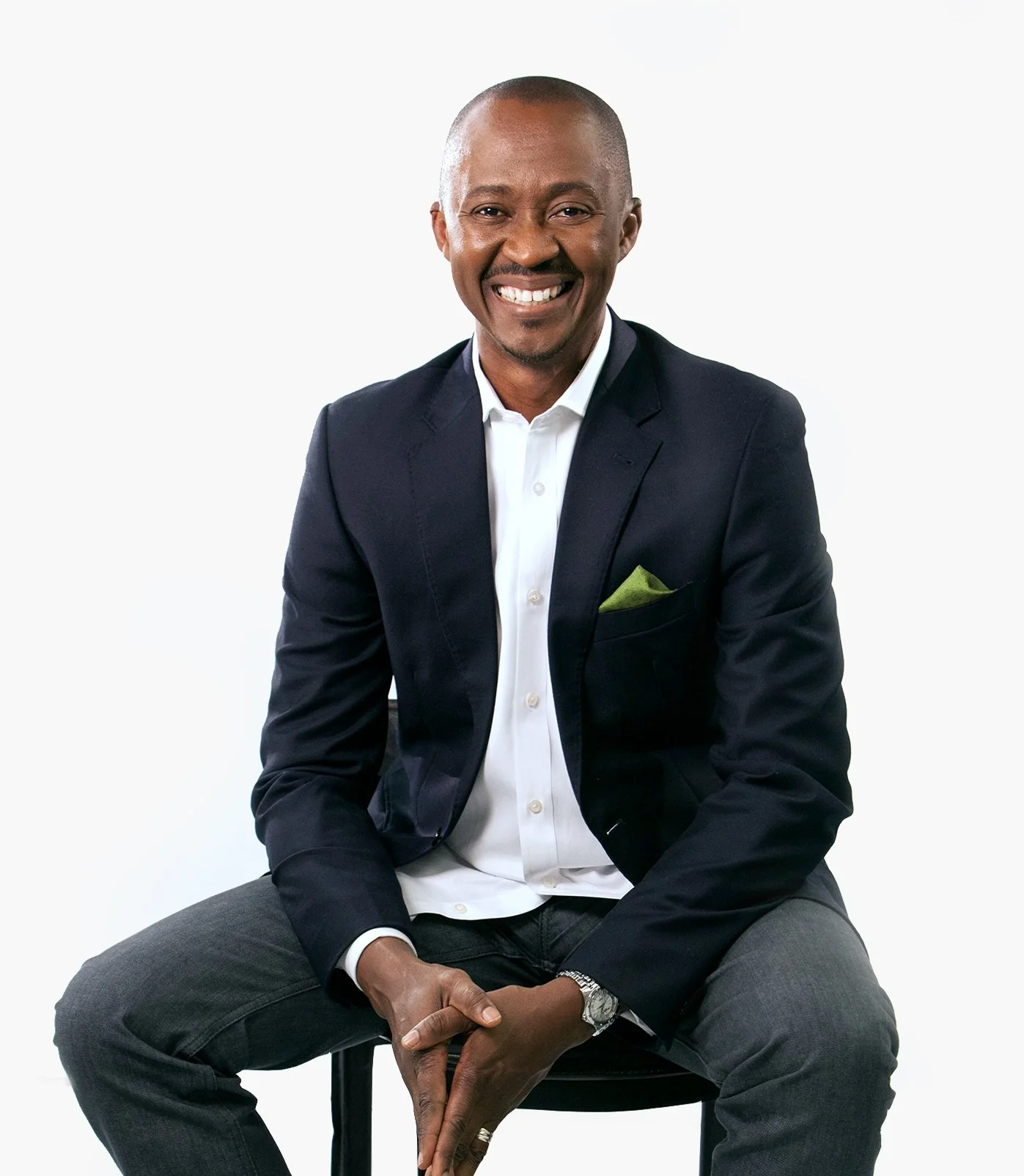 Portrait of Career and Leadership Coach, Kene Iloenyosi,  smiling in a black suit and white shirt, sitting on a stool with a white background.