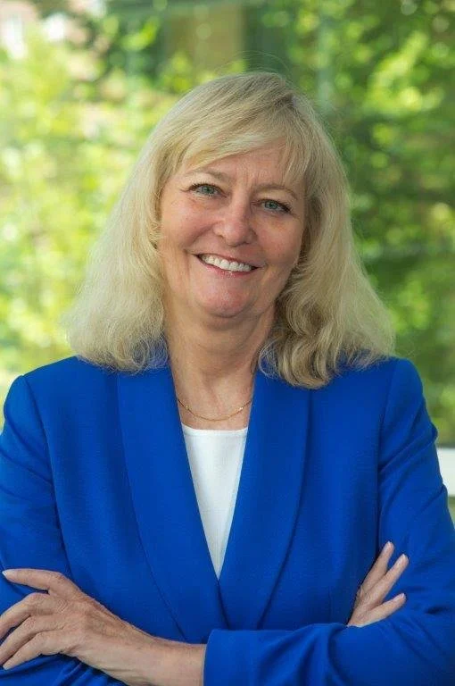 Middle-aged woman with blonde hair smiling, wearing a blue blazer and white top, standing outdoors with green trees in the background.