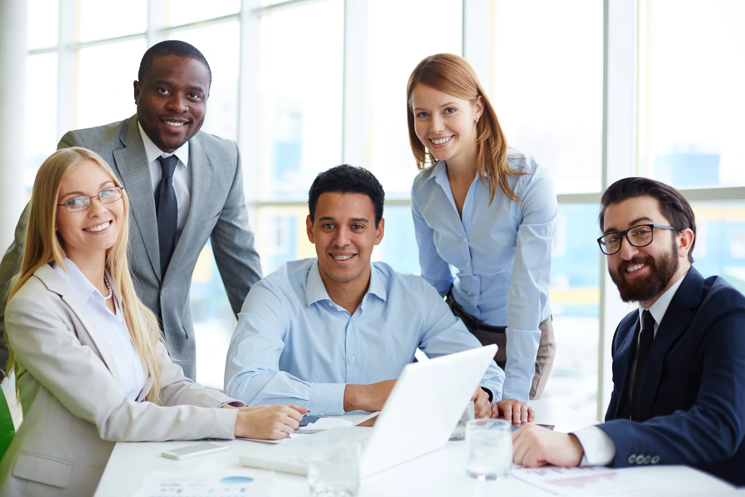 Group of six diverse business professionals in a career coaching meeting gathered in a modern office, some sitting and some standing, smiling and collaborating around a table with laptops and documents.