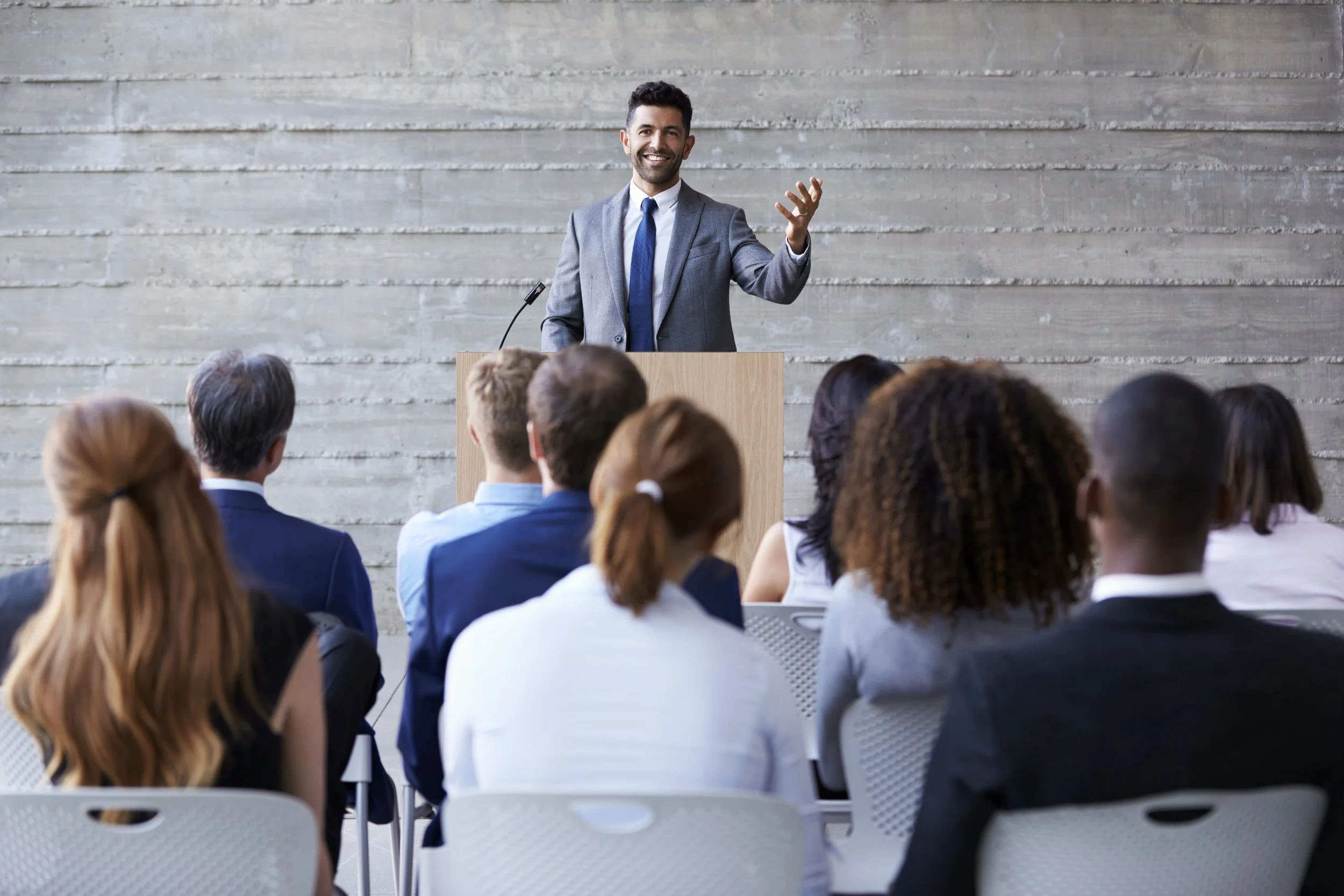 A man in a gray suit and blue tie speaking at a podium in front of an audience at a leadership workshop in a modern conference room with a concrete wall backdrop.