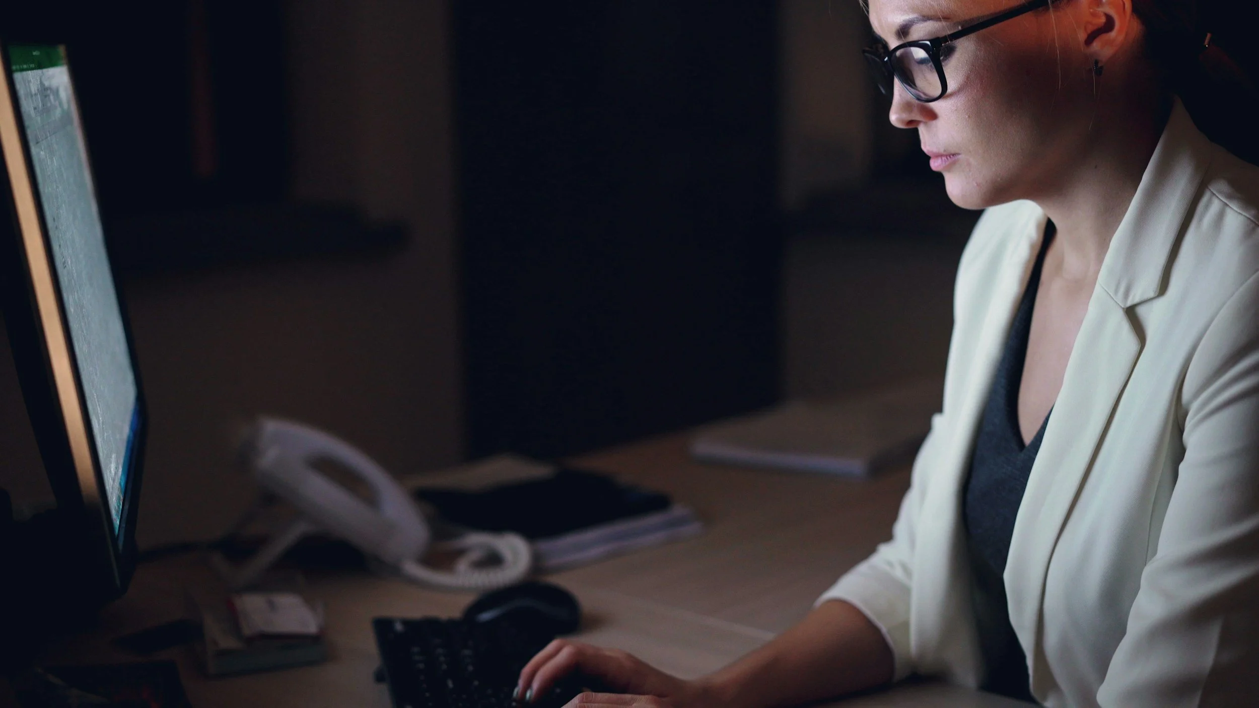 A woman with glasses working on a computer in a dimly lit office.