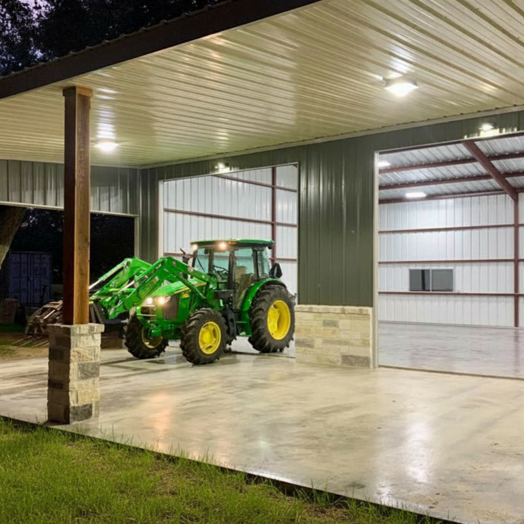 A green tractor parked under a Lean-to that is part of an enclosed metal building shop with concrete floor and wood and stone columns. Concrete has been clear sealed and shop has electrical package with LED lighting. Built in Cuero Texas