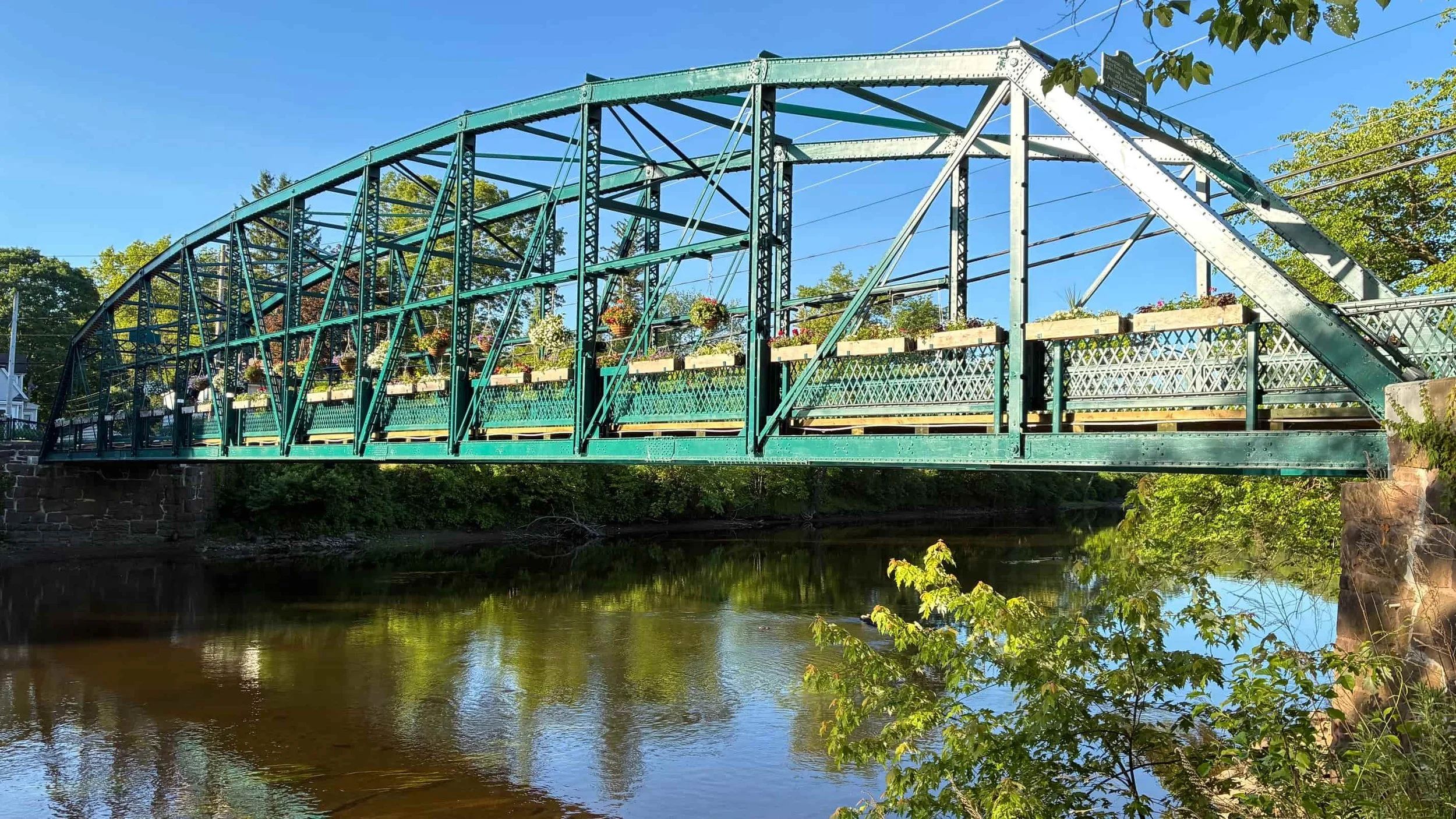 Green metal bridge over a river, with hanging flower baskets and trees on the sides.