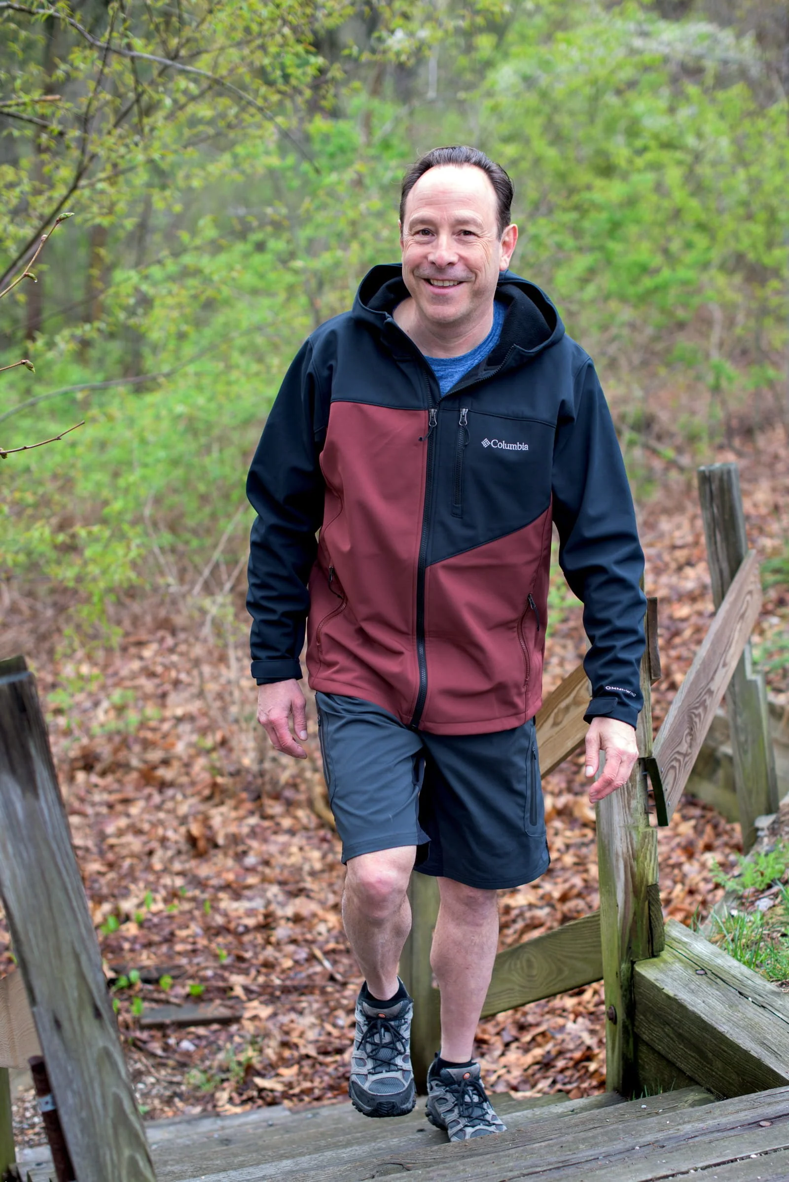A man walking on a trail in a wooded area, wearing a Columbia jacket, shorts, and hiking shoes.