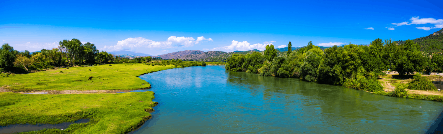 Scenic view of a river flowing through a lush green landscape with trees and mountains in the background under a blue sky with some clouds.