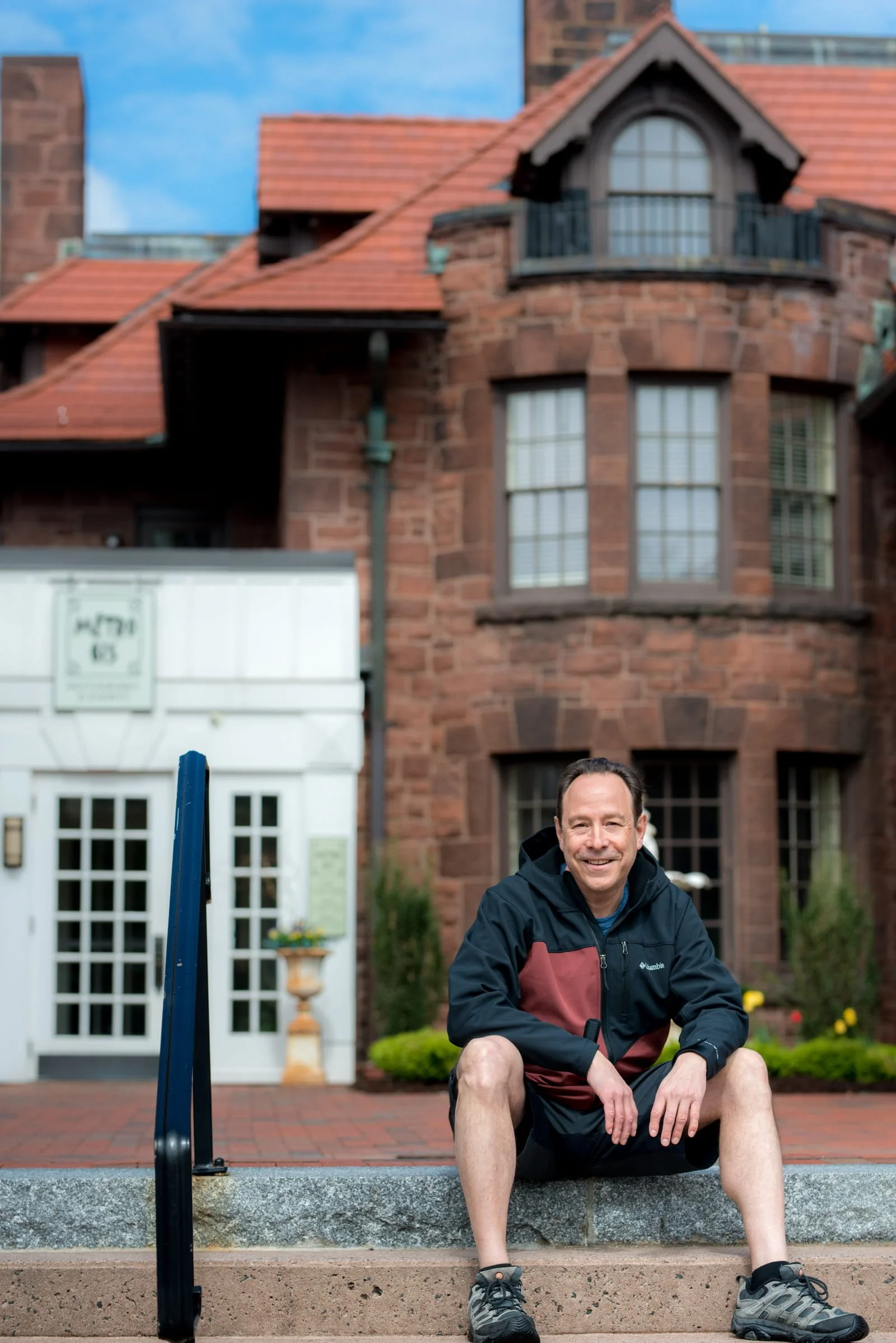 A man kneeling on the steps outside a brick house, smiling, with a blue sky overhead. There is a white building with a decorative door in the background, along with a garden and some potted plants.