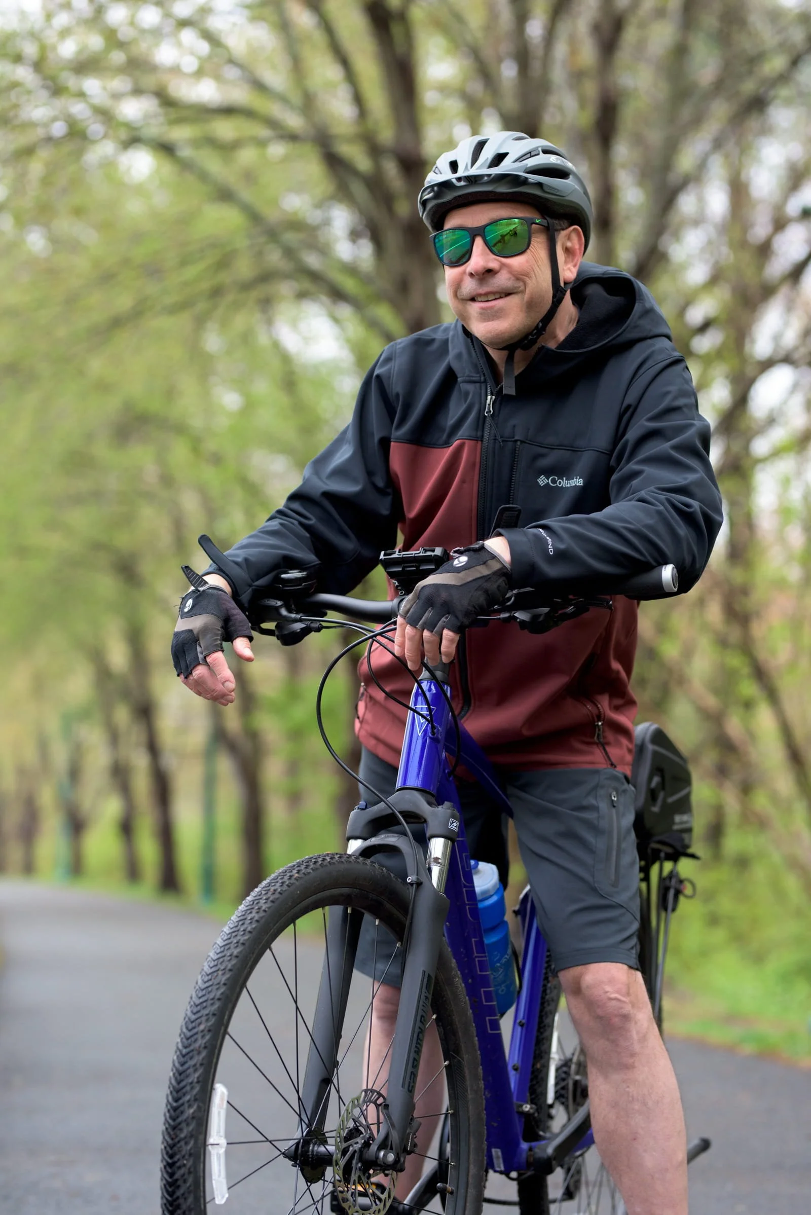 Man wearing a helmet, sunglasses, and a Columbia jacket riding a mountain bike in a wooded area.