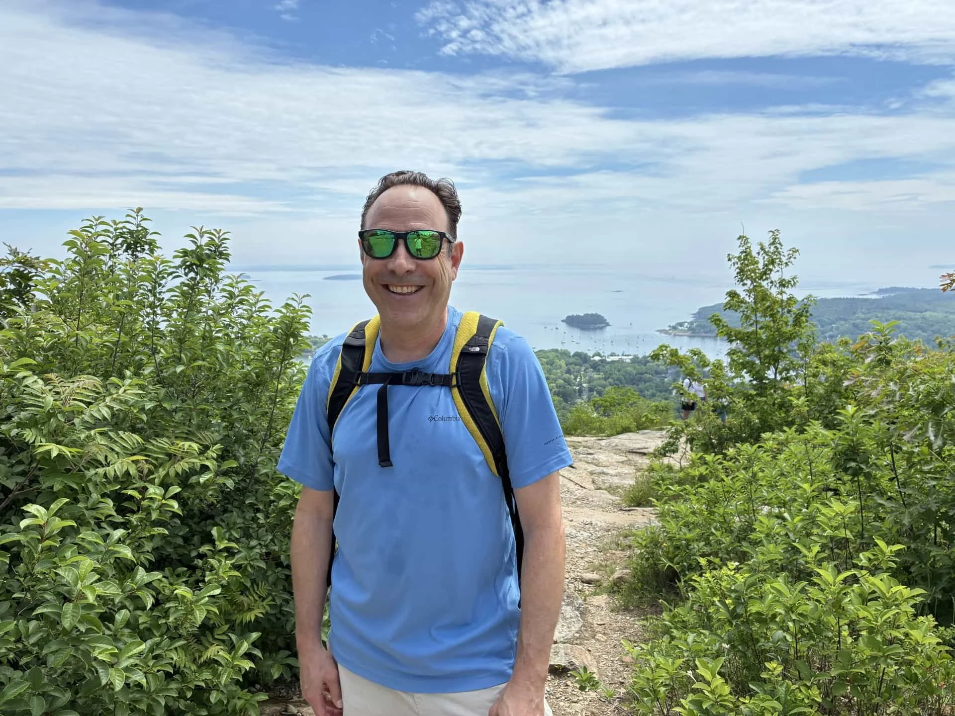 A smiling man wearing glasses and a blue Columbia t-shirt stands outdoors on a trail with greenery, overlooking a body of water with small islands in the distance under a partly cloudy sky.