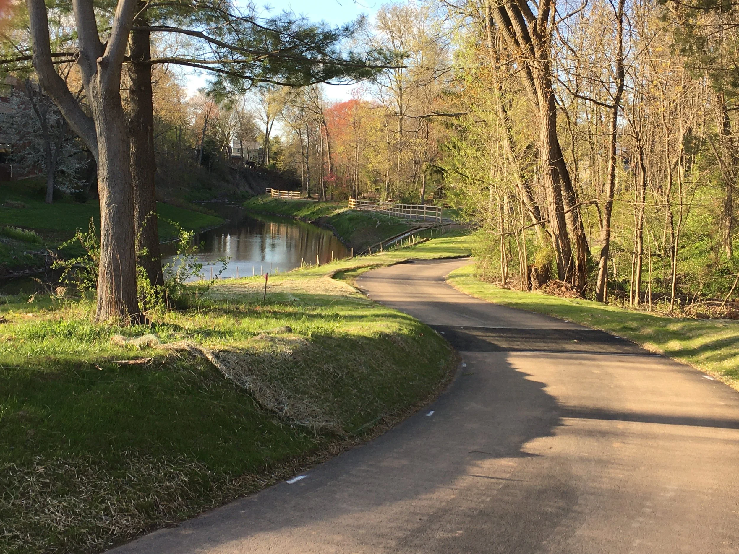 A scenic outdoor view with a paved walking path next to a small river or creek, lined with green grass and tall trees with budding leaves, suggesting springtime. The sky is clear and blue.