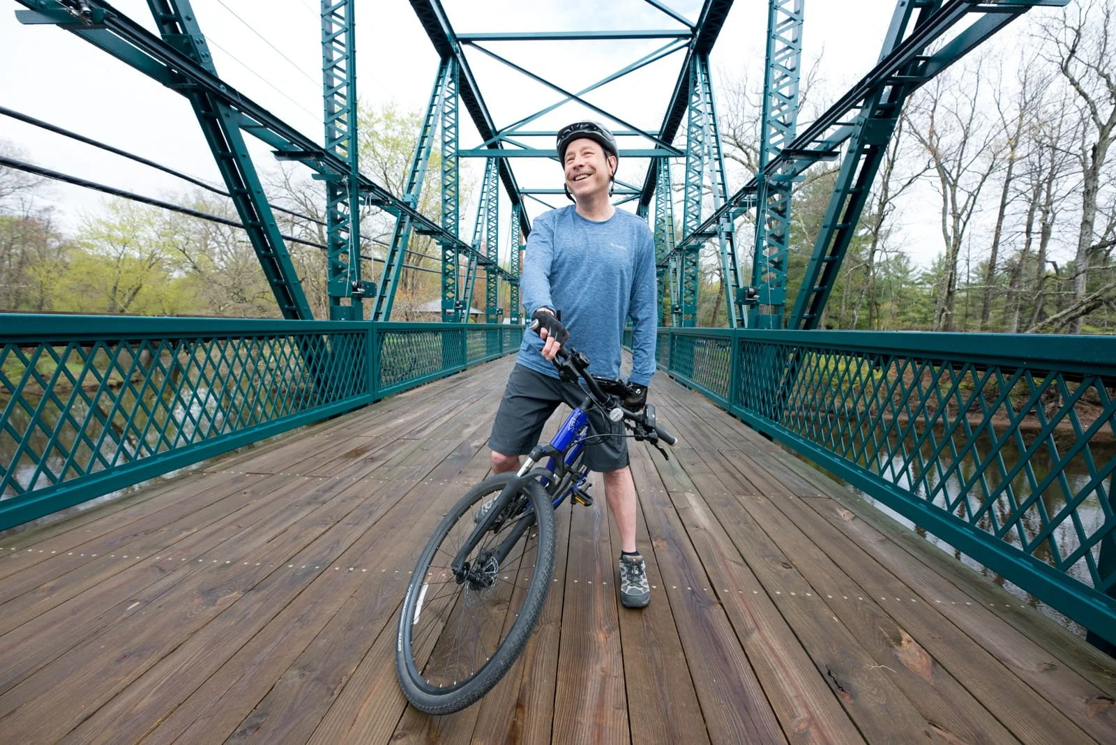 A man standing with a mountain bike on a wooden footbridge, smiling, wearing a helmet, gloves, blue long-sleeve shirt, and shorts, surrounded by trees.