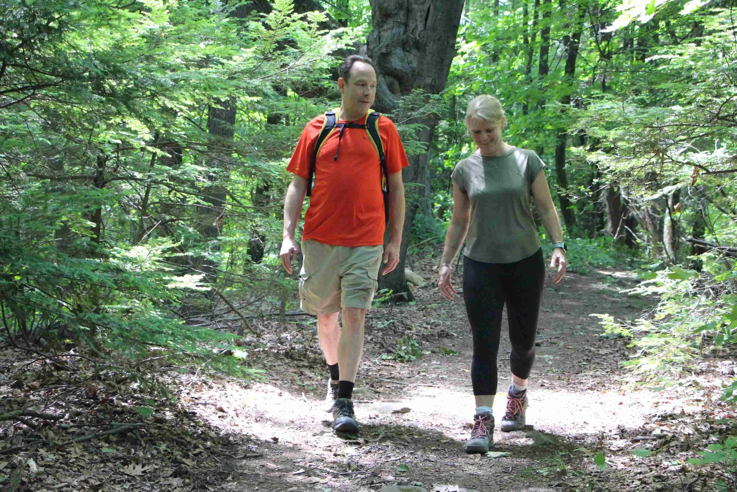 A man and woman hiking on a forest trail surrounded by lush green trees and foliage.