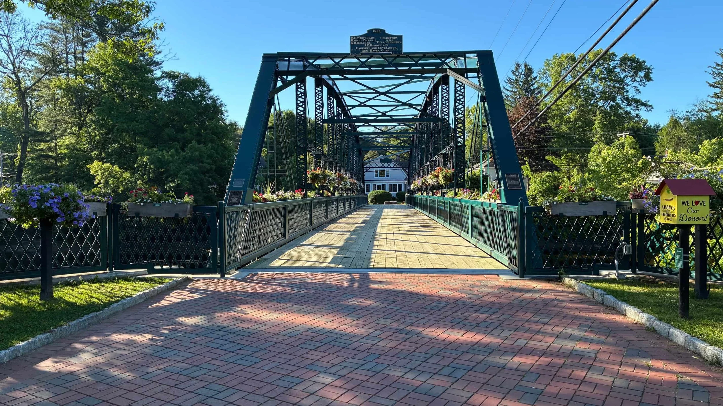 A pedestrian bridge with metal framing and hanging flower baskets, leading to a house surrounded by greenery. The scene is set on a sunny day with a clear blue sky.