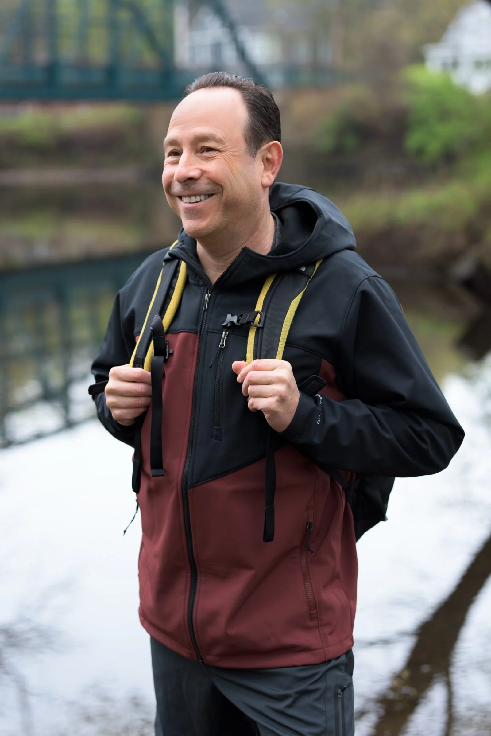 A man smiling outdoors near a river, wearing a black and maroon outdoor jacket, and carrying a yellow backpack strap.